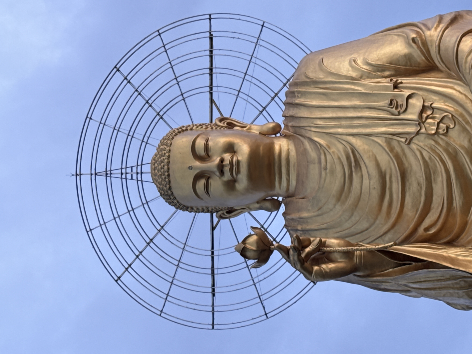 A golden Buddha statue, viewed from below, holds a flower. It is framed by a circular metal grid structure against a light blue sky. The statue appears to be located in Lâm Viên - Đà Lạt, Vietnam.