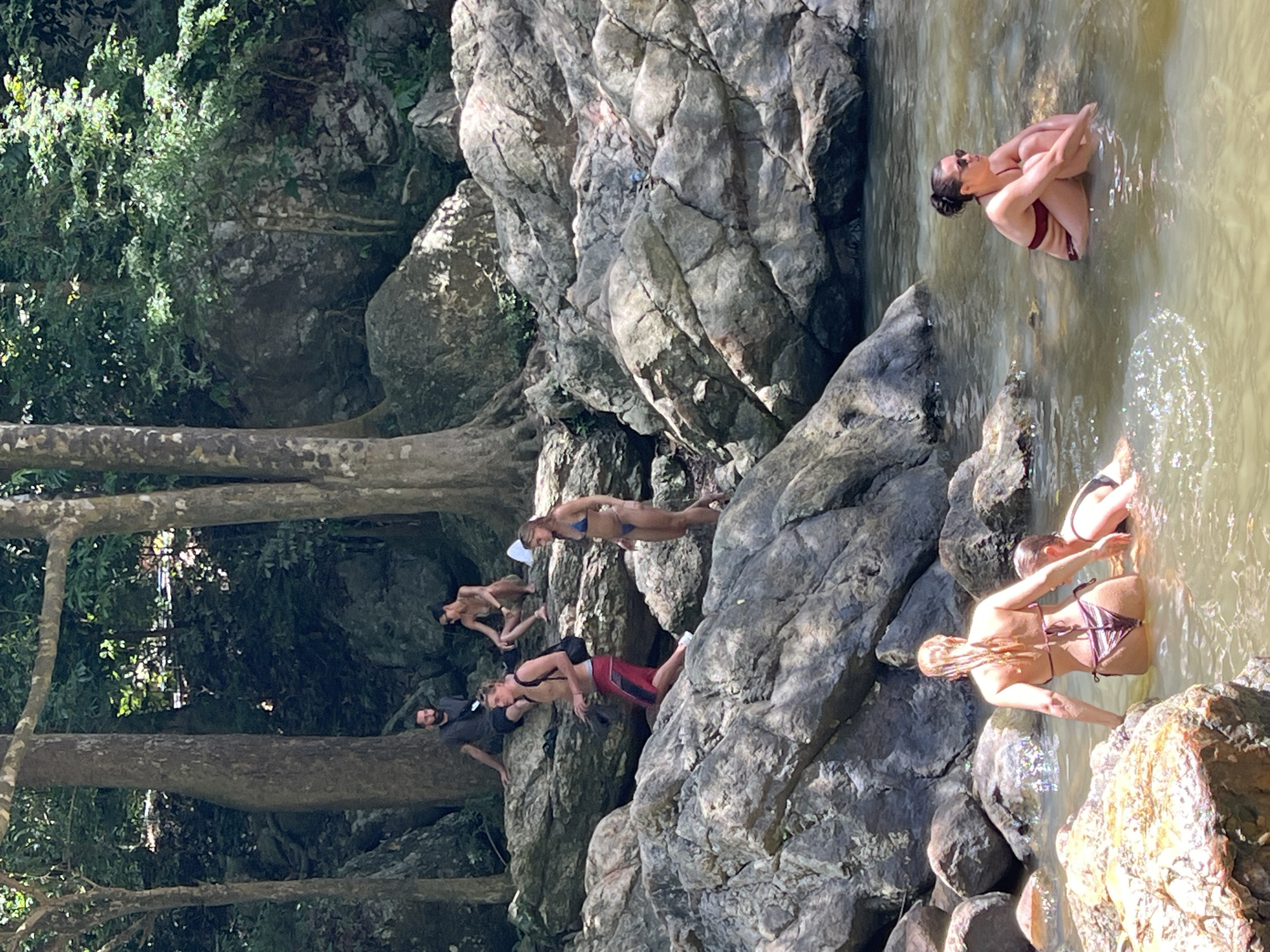 A group of people are gathered at a rocky waterfall in Baan Thurian, Thailand. Several individuals are in the shallow water, with some sitting on rocks and others wading. Two women in bikinis are in the foreground, one with her back to the camera and the other with her arms around a person partially submerged in the water. A man in red swimming trunks and a backpack is ascending the rocks, while another man and woman sit on a higher ledge. The environment is lush and green, with large trees and moss-covered rocks surrounding the waterfall. The water is murky, and sunlight filters through the trees, creating dappled patterns on the rocks and water.