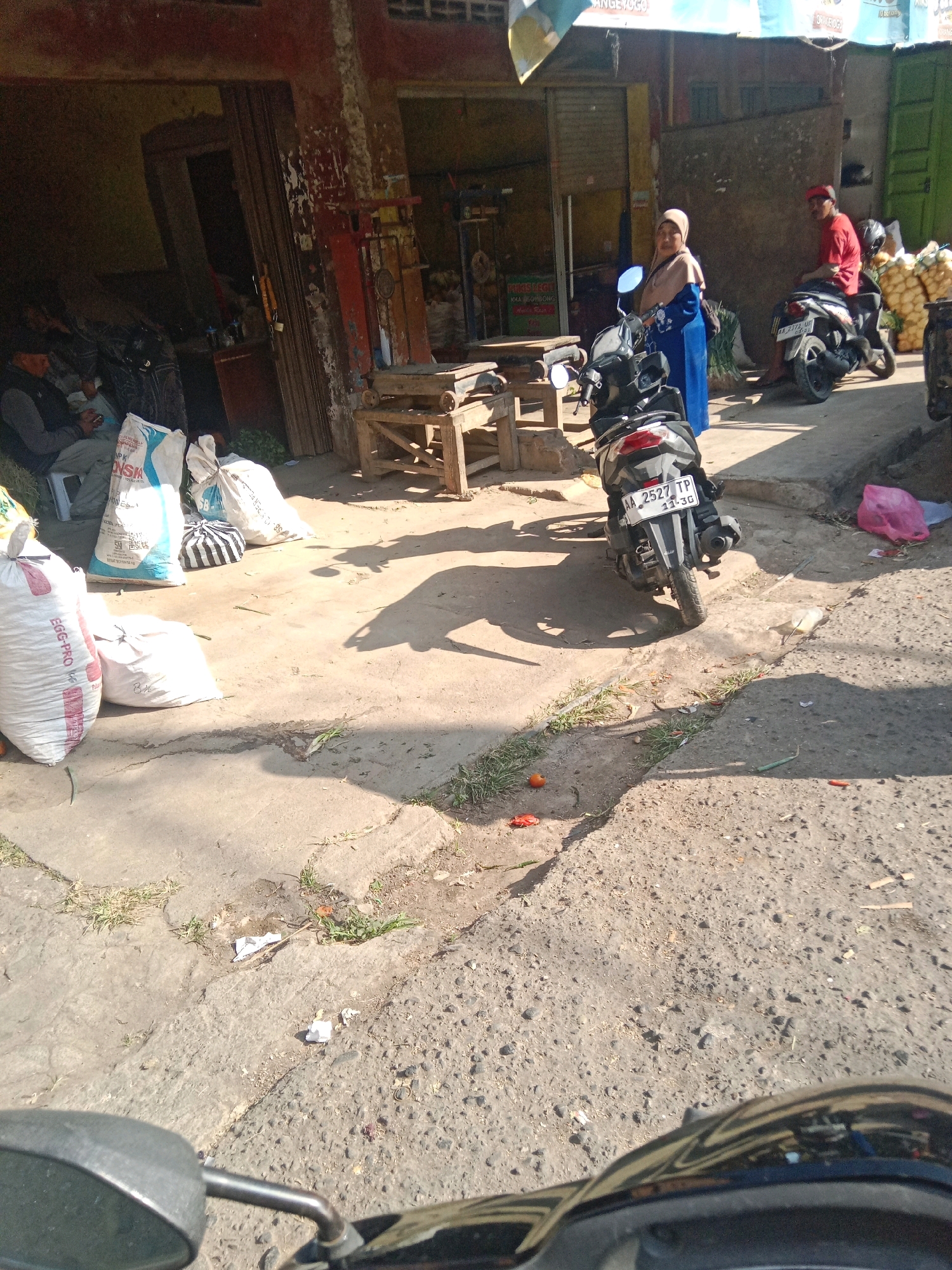 The image depicts a street scene in the city of Kertek, Indonesia, likely a market or roadside stall area. In the foreground, the handlebars and a rearview mirror of a motorcycle are visible, suggesting the photo was taken from a vehicle.
The midground reveals a woman wearing a blue hijab and a long blue dress, standing next to a parked motorcycle. To her right, a man in a red shirt and a red cap is seated on another motorcycle. Several large sacks, appearing to contain goods like grains or produce, are scattered on the ground. One sack, prominently displayed, has red lettering and the text "EGG-PRO". Another sack shows the brand name "ONSK".
The background shows a building with a reddish-brown facade, partially obscured by a green structure. The overall environment appears to be outdoors, with a concrete or asphalt ground surface. Shadows indicate it is daytime, and the lighting suggests bright sunlight. The scene has a somewhat unkempt appearance, with some debris and vegetation visible on the ground.