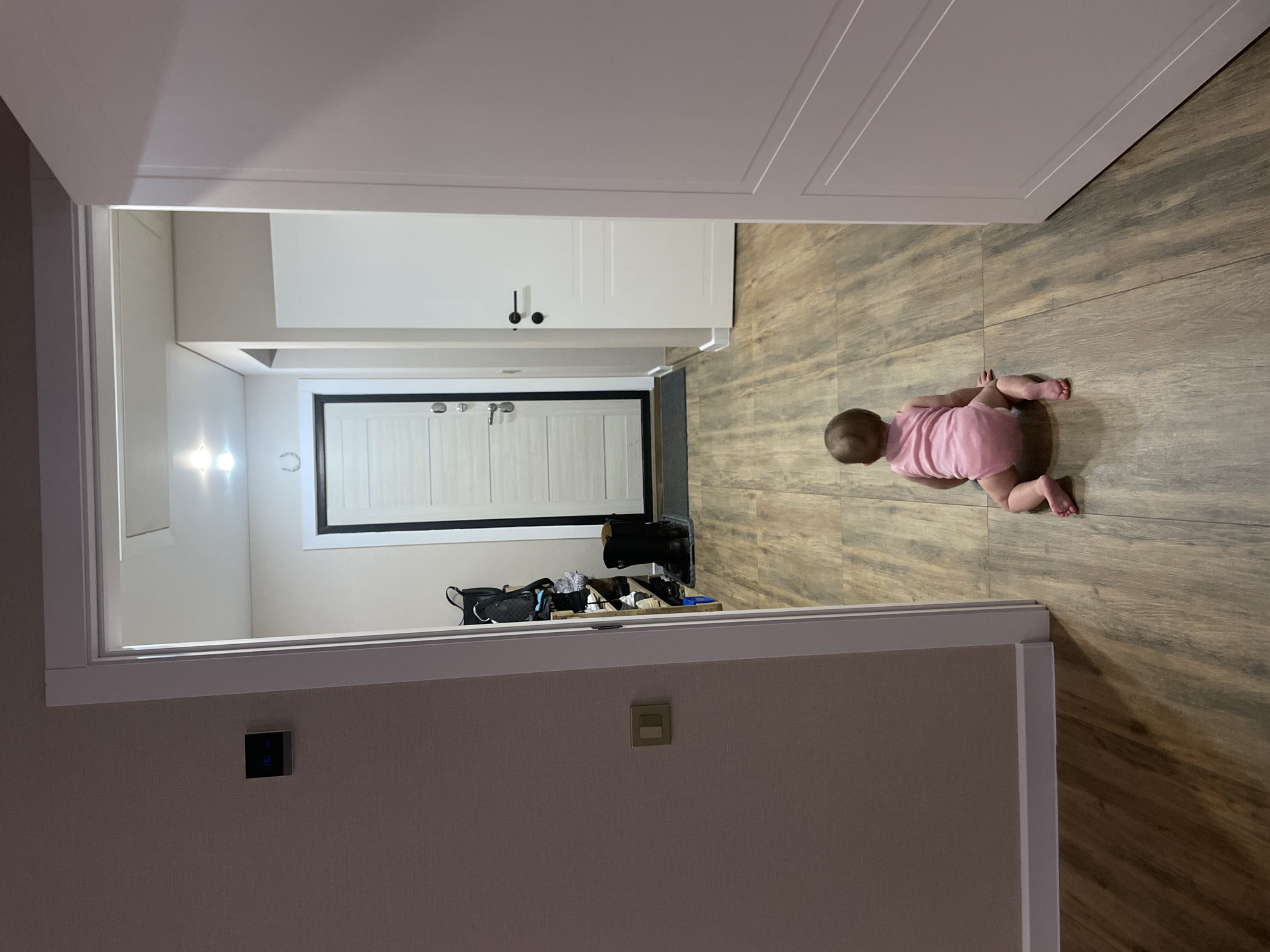 The image captures an overhead perspective of a domestic hallway, featuring a baby crawling towards the main entrance. The floor is covered with wood-look tiles or laminate in various shades of brown and gray.
In the foreground, a baby, dressed in a pink onesie, is seen from behind, crawling on all fours, moving away from the viewer. The immediate surrounding walls appear light-colored, possibly cream or light beige, with a modern light switch or smart home control panel with blue indicators visible on the left wall. Another standard light switch is on the right wall. The image is framed by a wide white doorway or archway.
Further down the hallway, the walls transition to white. Two bright, wall-mounted sconces illuminate the left side of the hallway. Above them, a small, metallic horseshoe-shaped decorative item is affixed to the wall. The hallway culminates at a white entrance door, distinctly outlined with a black frame and equipped with silver-colored hardware, including a handle and possibly a deadbolt. To the right of the door, a dark-colored storage unit or bench holds various items, including a black bag with a gold chain strap and other personal belongings. The ceiling is white, with some decorative paneling visible in the upper sections of the image.
The scene suggests a modern interior in Podshivalovo, Russia. The artificial lighting indicates it might be evening or an area lacking natural light, though no windows to the exterior are visible. No discernible text is visible in the image.