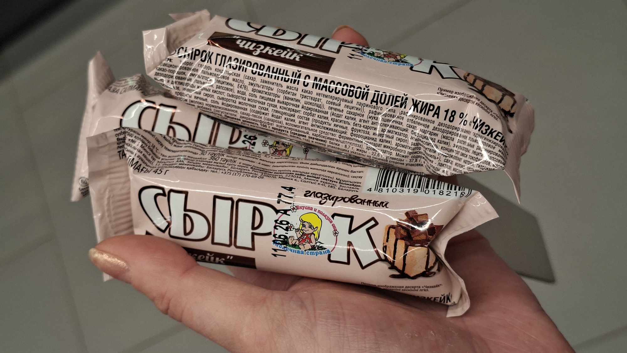 A close-up shot captures a hand holding two packages of "Сырк" ice cream bars, one in front of the other, against a muted background. The ice cream bars are pink and white with Cyrillic text and an illustration of a girl. The left bar features the text "СЫРК" in large, bold letters. A date "11.06.26 A 77.4" is visible, along with a smaller circular illustration of a blonde girl with flowers. The right bar, partially obscured, also shows the "СЫРК" branding. Ingredients and nutritional information are printed in smaller Cyrillic text on the packaging. The background appears to be an indoor setting with a smooth, light-colored surface. The location is identified as Smalyavichy, Belarus.