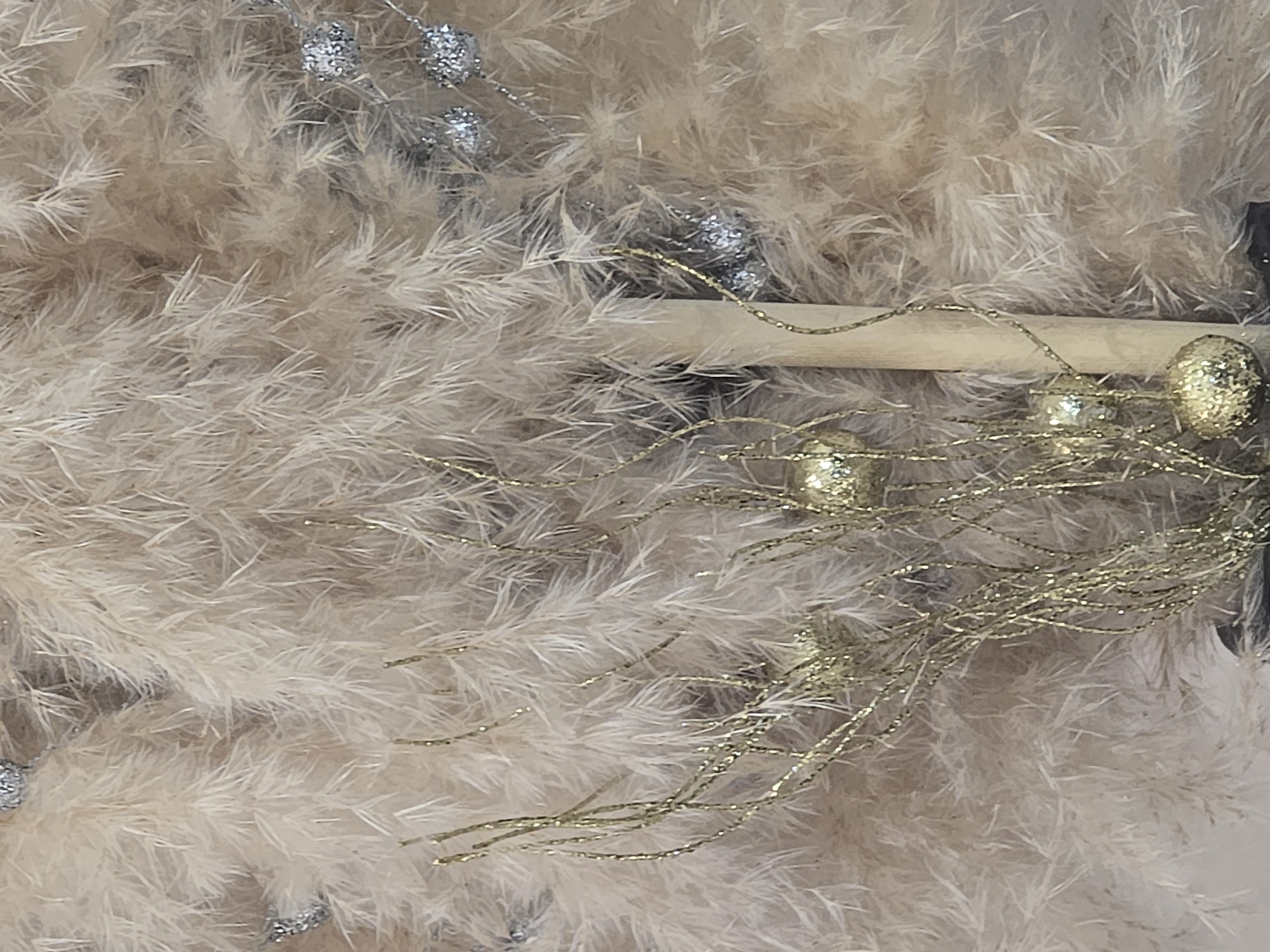 The image is a close-up of a decorative arrangement featuring light beige, fluffy reeds or grasses interspersed with shimmering silver and gold accents. The gold elements appear to be thin, twisted branches adorned with small, glitter-covered spheres. A vertical, light-colored wooden or bamboo rod is partially visible on the right side of the frame. The background is entirely filled with the soft, feathery texture of the reeds, creating a bokeh effect that blurs any distant details. The lighting is soft and diffused, highlighting the textures and subtle sheen of the decorations. There are no people or animals visible in the image, nor any discernible signs of a specific location within the city of Ulan-Ude, Russia. The image focuses solely on the decorative composition itself.