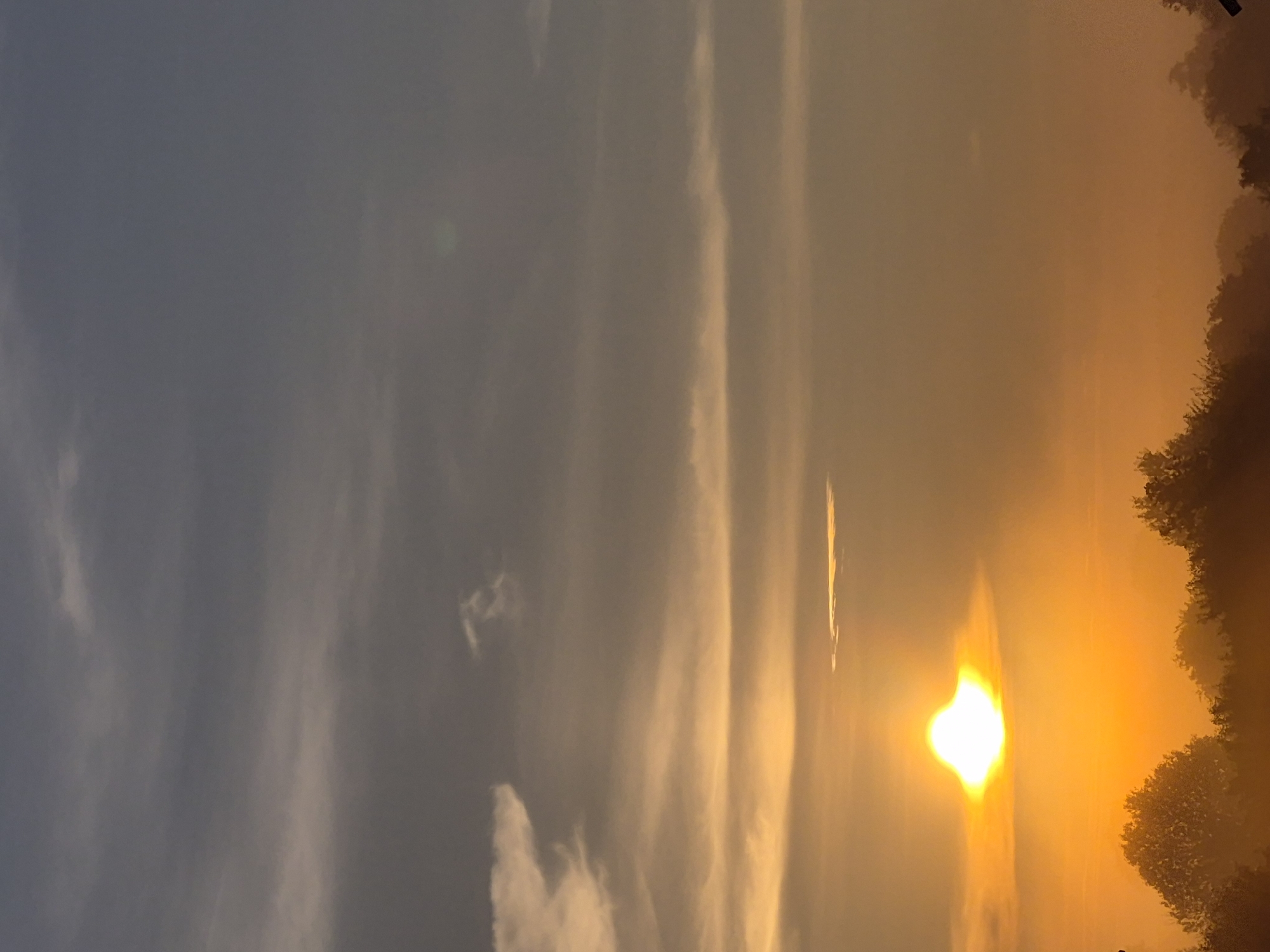This is a photograph taken during sunrise in Sooblase, Estonia. The sun is a bright, glowing orb near the lower left of the frame, casting an intense orange and yellow light across the sky. The sky is filled with wispy clouds, some appearing white and others tinged with the warm hues of the sunrise. Below the sky, a silhouetted treeline marks the horizon, with the tops of the trees dark against the luminous sky. The overall impression is one of a tranquil and serene dawn in a natural landscape.