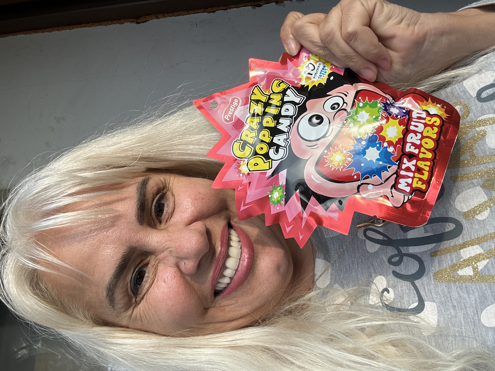 A woman with fair skin and long, blonde hair is holding a package of "Crazy Popping Candy Mix Fruit Flavors" in her right hand. She is smiling and looking at the camera. She has a nose piercing and is wearing a grey t-shirt with a gold and black design. The background appears to be a light grey wall or surface. There is nothing to indicate this image was taken in Sofia, Bulgaria.