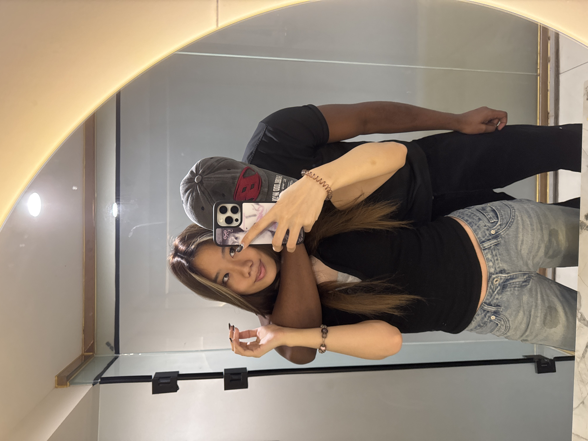 A couple is posing in a bathroom mirror for a selfie. The woman is in the foreground, holding a phone with a patterned case. She's wearing a black shirt, light-wash jeans, and has highlighted hair. The man, behind her, is wearing a black shirt and black pants with a cap. The mirror has a rounded top and is surrounded by a light frame. The background includes tiled walls and a shelf. There are no indicators of this image being taken in Yuzhong District, China.