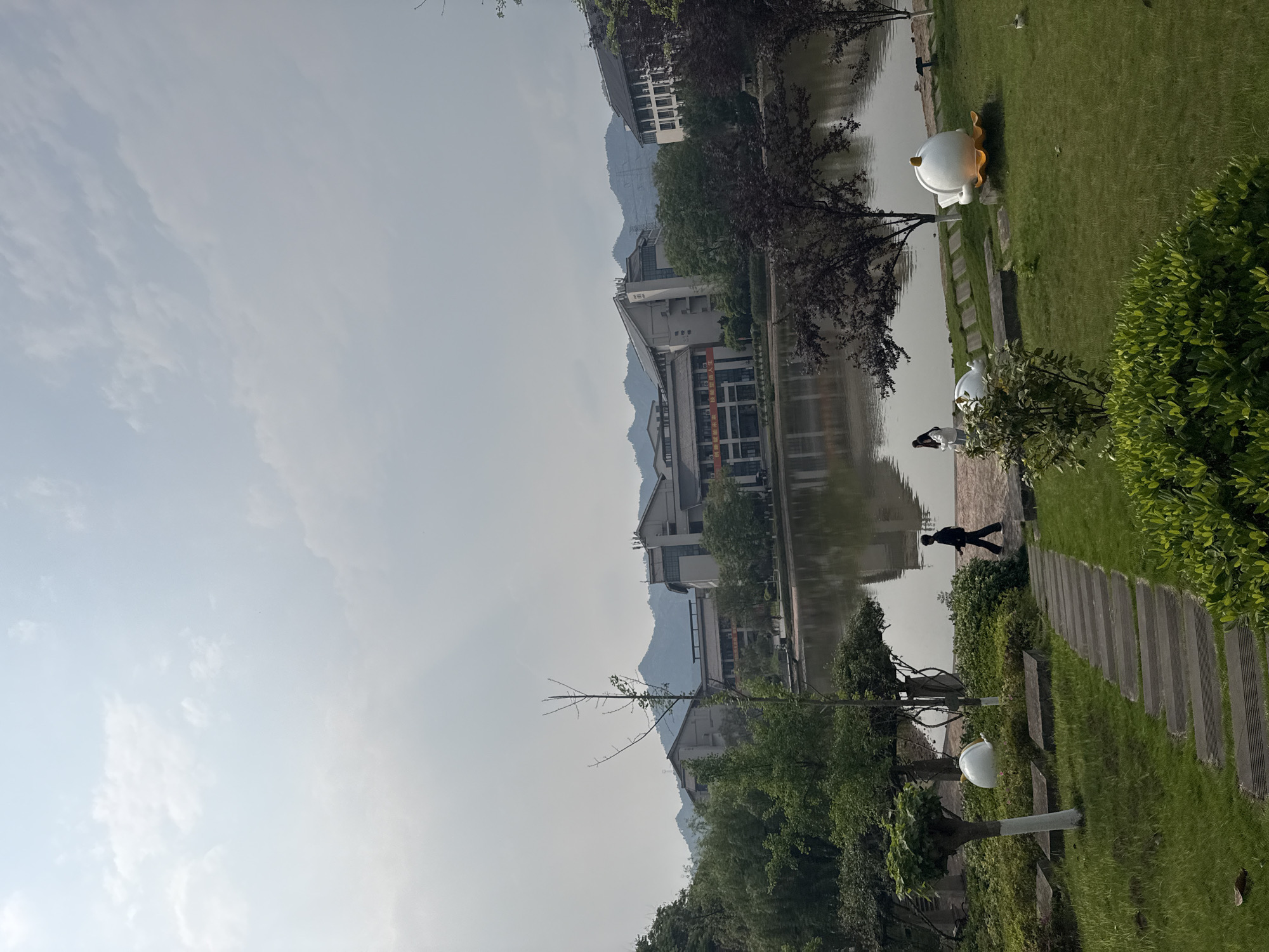 The image depicts a scene in Jiangjin, China, alongside a body of water, possibly a lake or river, reflecting buildings and trees. A person is walking along a paved path, while other individuals are visible near the water's edge. Sculptures resembling ducks or geese are situated on the grassy area next to the path. Mountains can be seen in the distance, creating a backdrop to the buildings. The sky is overcast, casting a diffused light over the scene. The environment includes trees, bushes, and manicured grass.