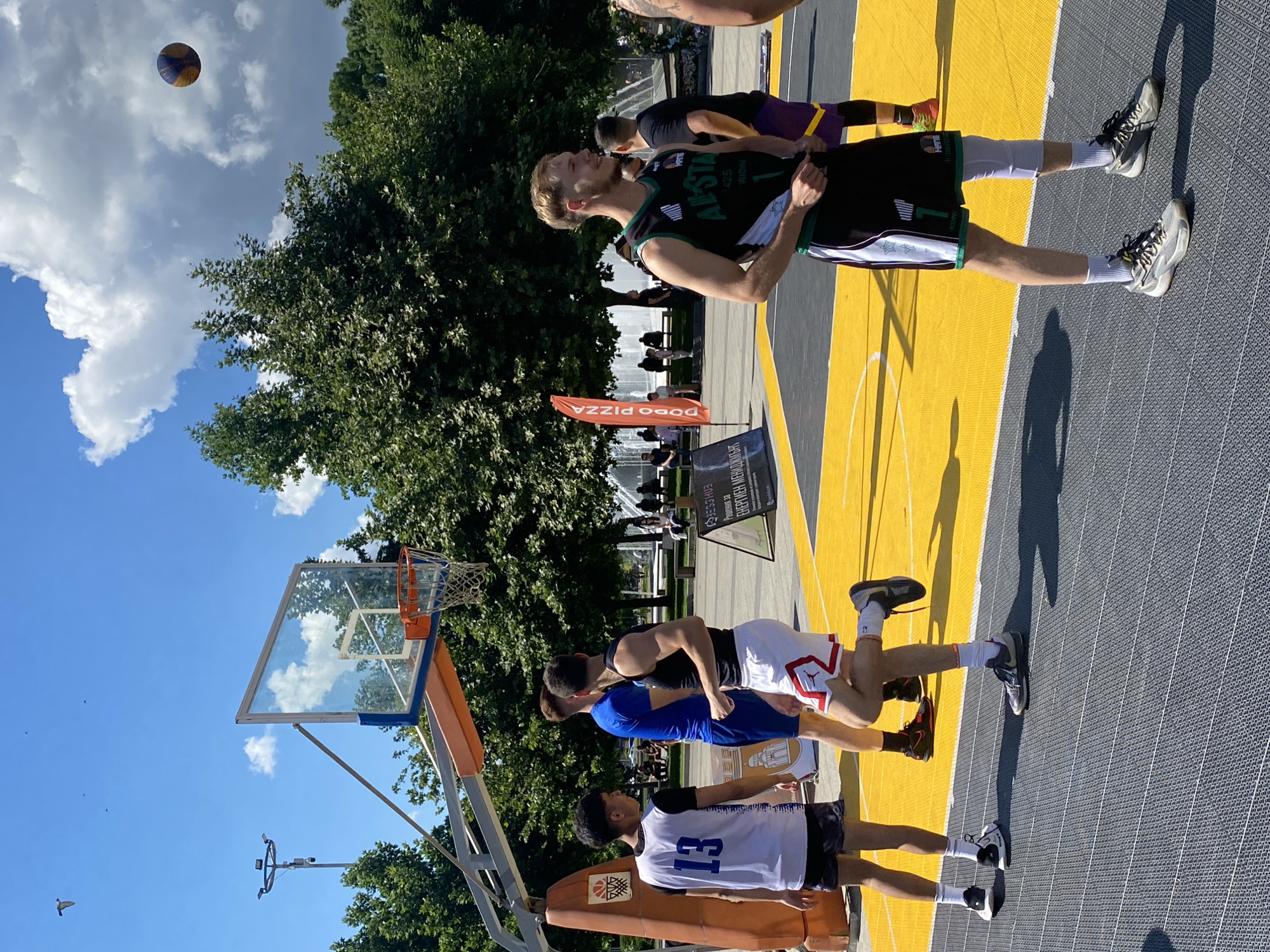 The image shows a basketball court in Pleven, Bulgaria, likely in the city center, given the presence of a fountain and trees commonly found in urban parks. The court has a bright yellow playing surface with grey surrounding areas. Several young men are present, dressed in athletic wear, engaged in what appears to be a basketball game or practice. One individual in the foreground wears a black and green basketball jersey with the number '1' and various logos, including "All Star." Another person to his left is wearing a white jersey with the number '13'. To the left, a basketball hoop with a glass backboard is visible. In the background, a fountain operates, and trees line the perimeter of the court. The sky is clear blue with some scattered clouds, and a basketball is visible in mid-air. The overall scene depicts a sunny day in an urban outdoor recreational space.