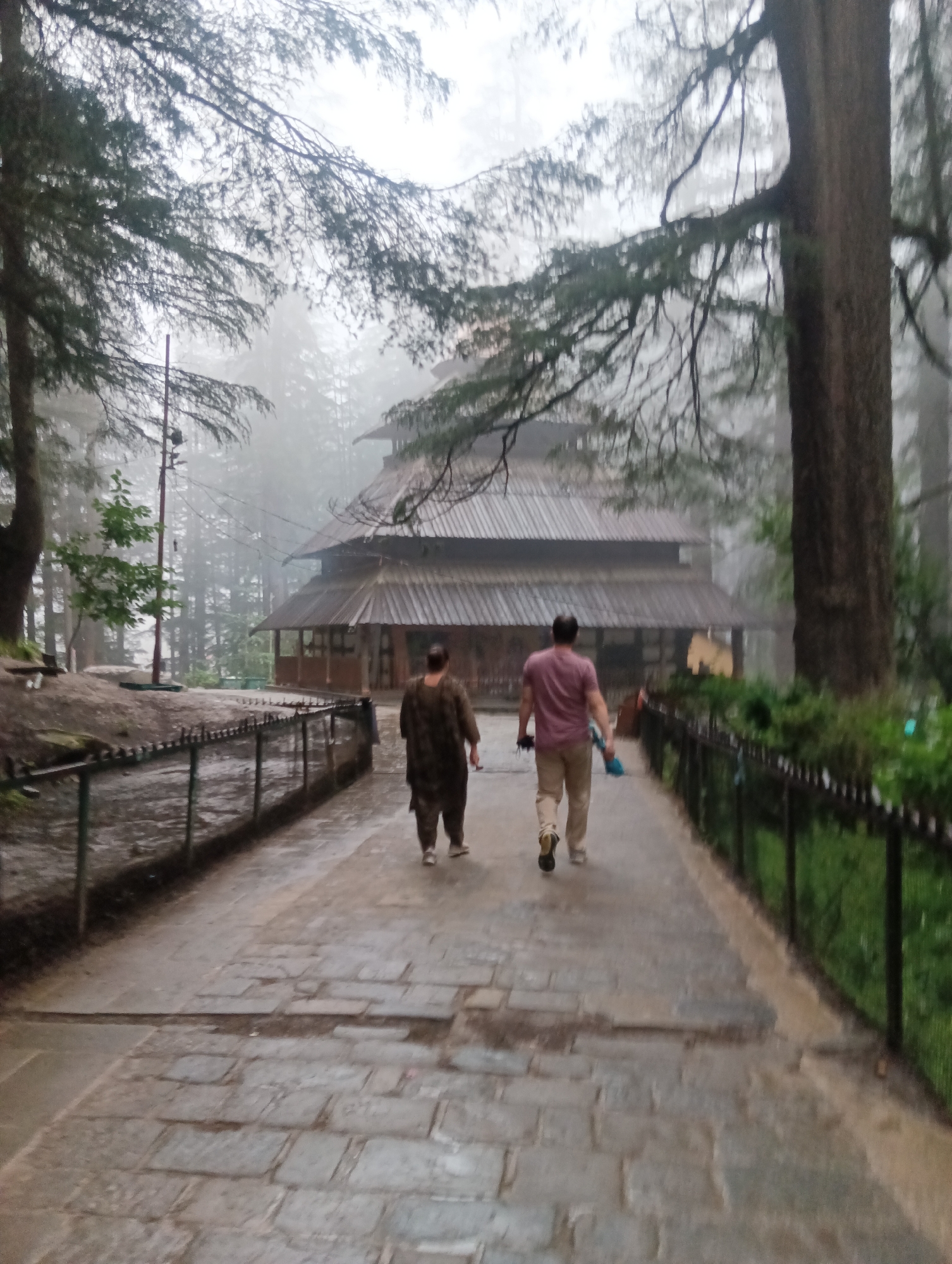 The image shows a foggy day at the Hadimba Devi Temple in Manali, India. Two individuals, one wearing a dark patterned outfit and the other a maroon t-shirt and beige pants, are walking away from the camera down a stone pathway. A traditional wooden temple with a multi-tiered, sloping roof is visible in the background, partially obscured by trees and mist. A dark metal fence runs alongside the pathway. Tall pine trees dominate the surrounding environment, their branches and foliage visible through the atmospheric haze. The ground appears damp, suggesting recent rain or the general dampness of a foggy forest.