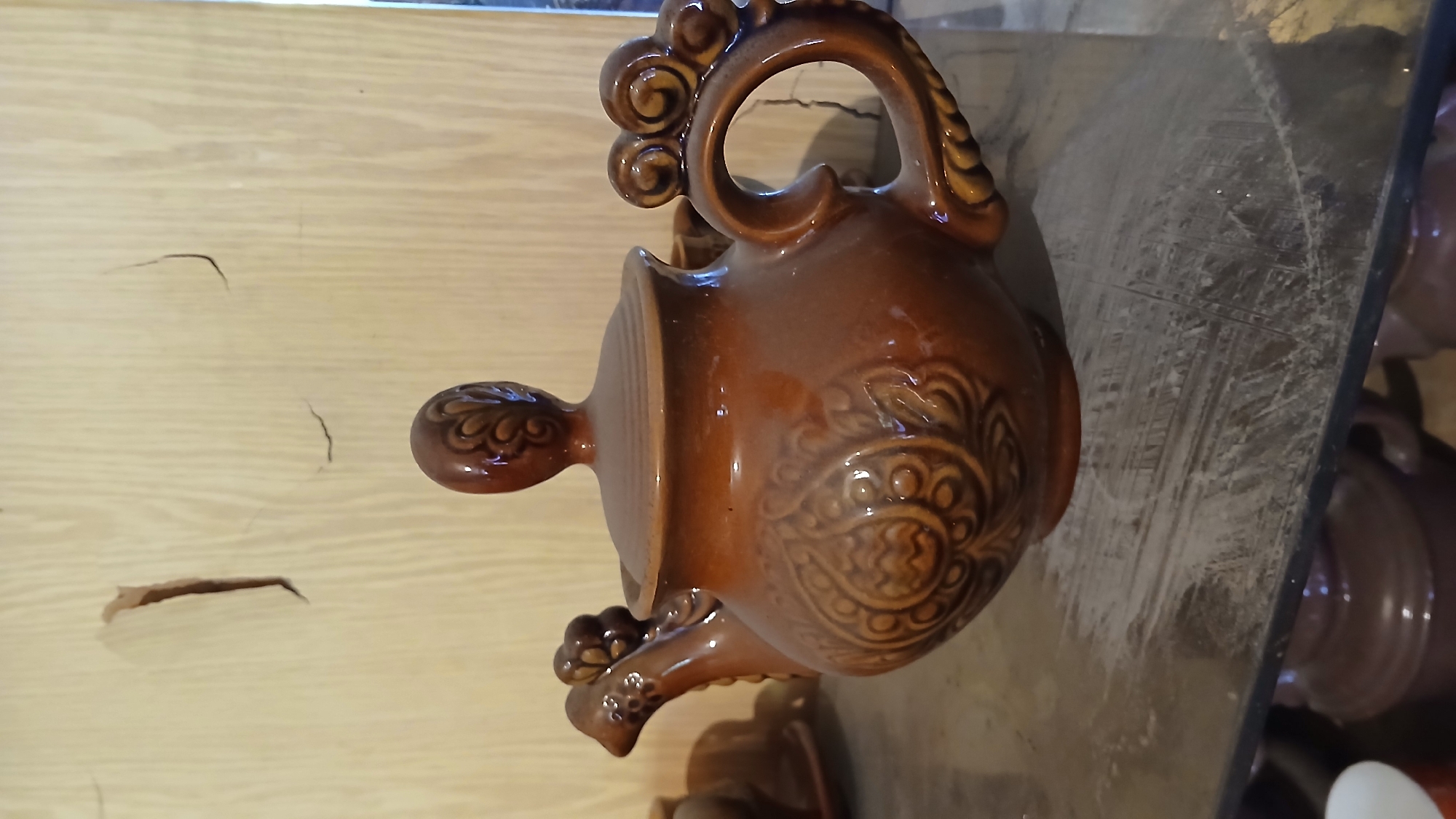 The image shows a brown ceramic pitcher with ornate detailing. It has a looped handle and a prominent spout. The pitcher sits on a reflective surface, possibly a countertop, with other similar items visible in the background. The surrounding environment includes a light-colored wooden wall. There is no information available to indicate the location is in Dnipro, Ukraine.
