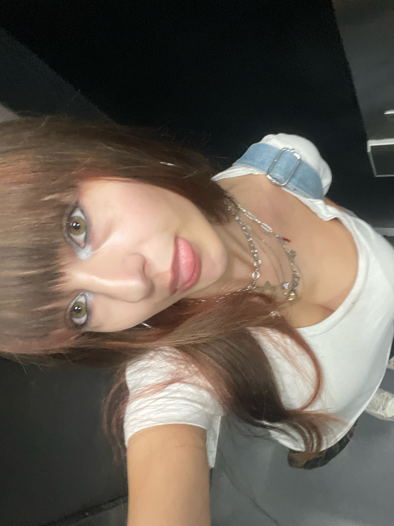 A young woman with brown hair and light green eyes is looking up at the camera. She is wearing a white top with a denim strap over her shoulder and several silver necklaces. Her makeup includes light blue eyeshadow. The image appears to be taken indoors, with dark walls in the background. There is no information to suggest this image was taken in Raanana, Israel.