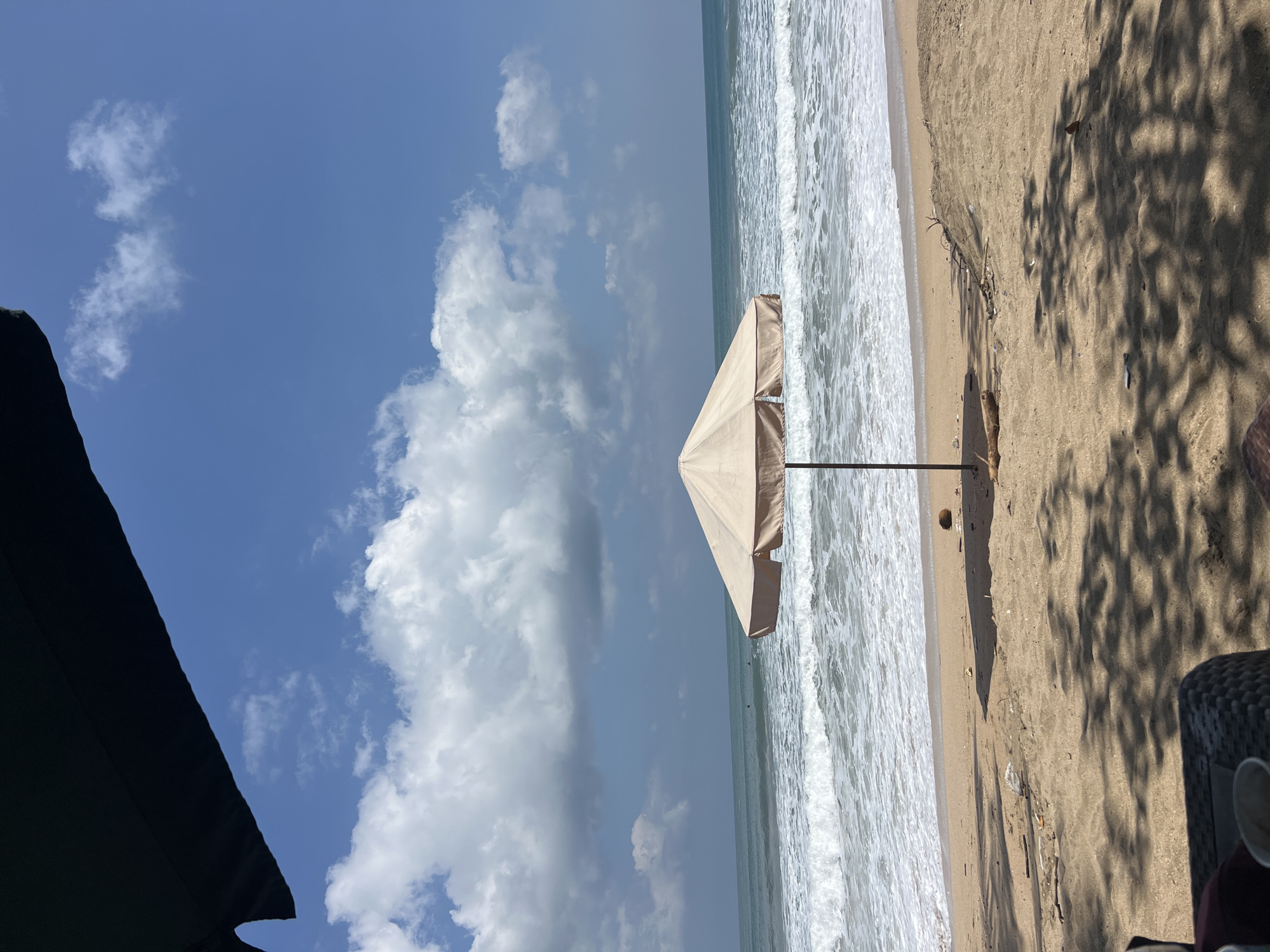 This is a photograph taken from a shaded area looking out towards the ocean on a beach in Kuta, Indonesia. A large, tan beach umbrella is positioned in the center of the frame, casting a shadow on the sand. The umbrella is supported by a dark pole. In the background, the ocean exhibits gentle waves with white foam, and the water is a deep blue-green. The sky above is a bright blue with scattered white cumulus clouds. The beach itself is made of light brown sand, with some debris and a coconut visible on the ground. The foreground is shaded, suggesting the viewer is seated under another umbrella or structure, with a wicker chair partially visible at the bottom. The overall scene depicts a tranquil beach environment.