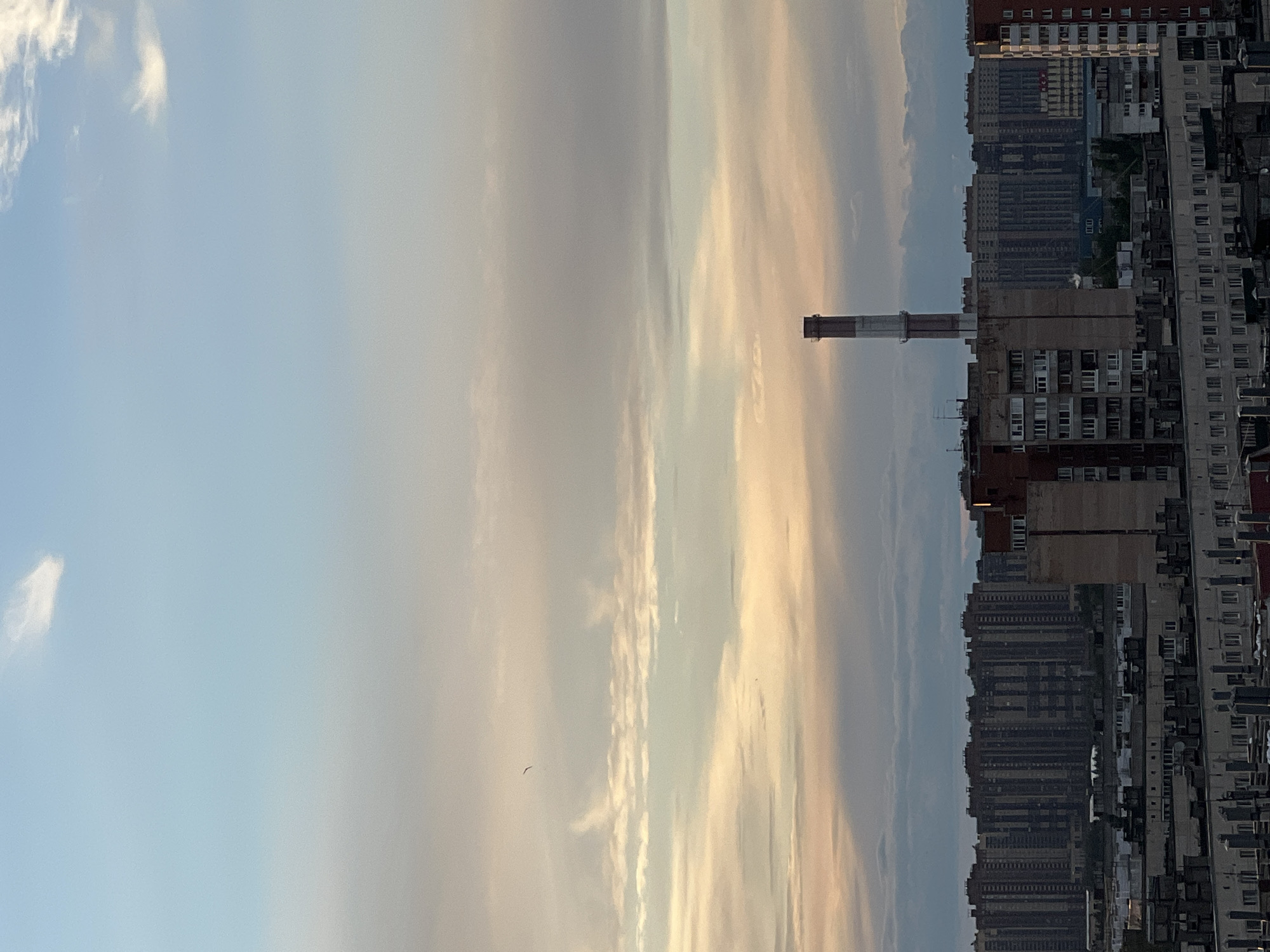 The media file is an image of an urban skyline in Sinyavino, Russia. The image is oriented vertically, showing a vast expanse of sky on the left and a dense arrangement of multi-story buildings on the right. The sky is a pale blue, transitioning to streaks of soft orange and yellow clouds, suggesting either sunrise or sunset. A solitary bird is visible flying in the sky. Dominating the cityscape is a tall, slender smokestack or industrial chimney with a band of red and white near its top.
The buildings are primarily apartment blocks, appearing to be of Soviet-era design, characterized by their uniform window patterns and concrete facades. Some of the buildings are taller and more modern, while others are older and appear more weathered. The density of the buildings creates a sense of a highly populated urban environment.
The time of day is likely early morning or late evening, given the soft, diffused light and the colors of the sky. The weather appears to be clear, with scattered clouds. There is no visible text in the image. The overall mood conveyed by the image is one of urban life under a serene sky, with a hint of industrial presence from the chimney.