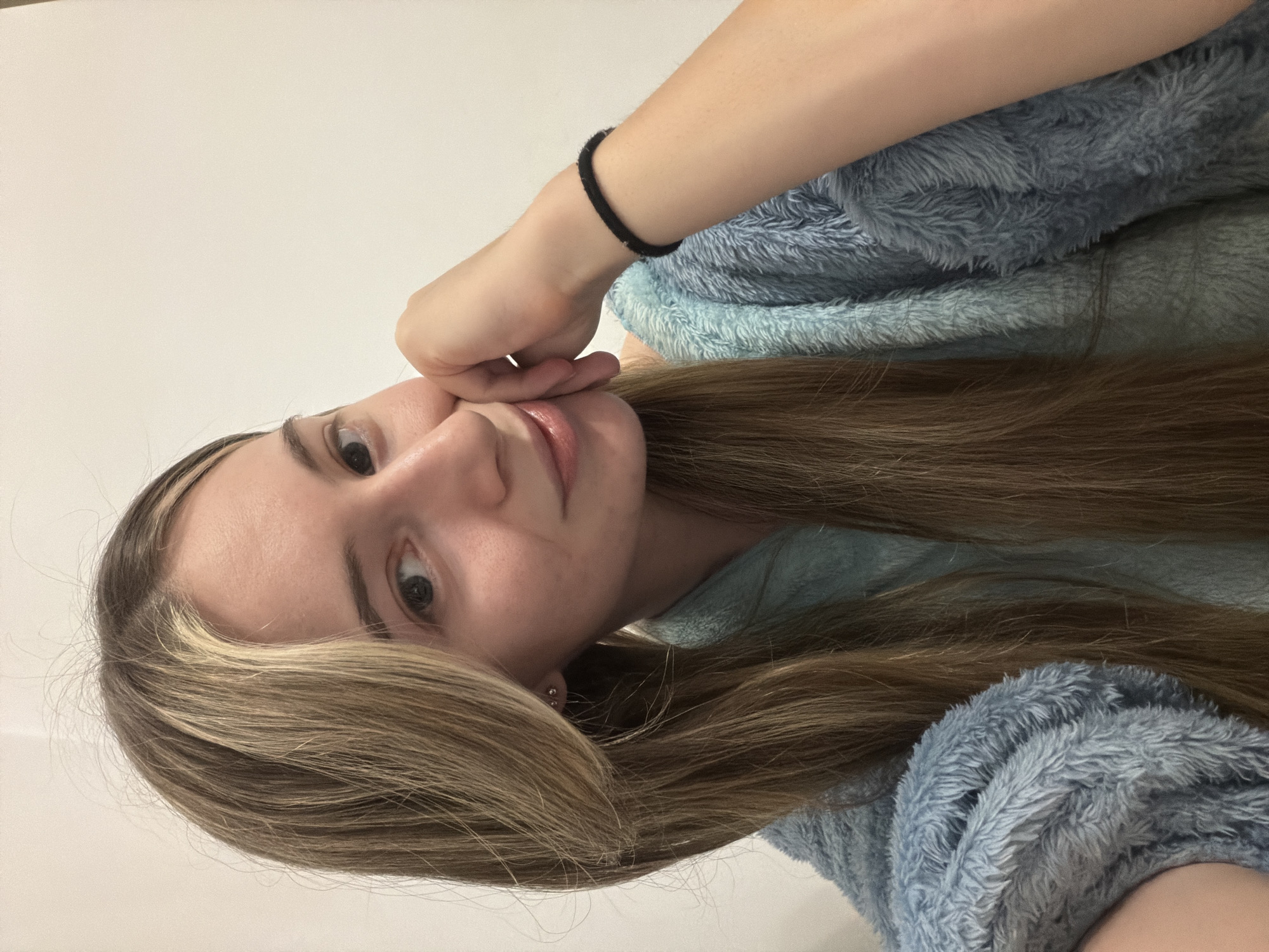 A woman is holding her face with her hand, wearing a light blue fuzzy robe. A black hair tie is on her wrist. Her hair is long and light brown with blonde highlights. The background is a plain white wall. There is no evidence to suggest the location is Plovdiv, Bulgaria.
