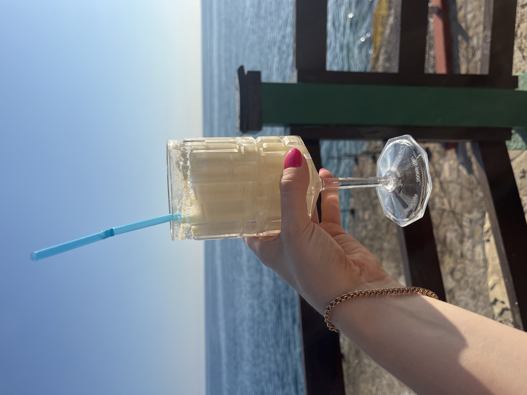 A person's hand, adorned with a rose gold bracelet and a bright pink fingernail, holds a clear plastic cup filled with a pale yellow, frothy beverage. A blue plastic straw extends from the drink and points towards the sky. The cup is octagonal and has a textured pattern on its surface. The hand is holding the stem of the cup, which is clear and also octagonal, with some faint text visible on it.
The background consists of a vast expanse of clear blue sky meeting the calm, deep blue sea. In the mid-ground, a dark wooden railing with a green horizontal bar creates a boundary. The sea is slightly textured with small waves and reflections of sunlight.
The overall impression is one of a relaxed and pleasant day, likely during daylight hours, given the bright sunlight and clear sky. The setting suggests a waterfront location, possibly a cafe or bar in Alanya, Turkey, overlooking the sea. The focus on the refreshing drink and the beautiful view evokes a sense of leisure and enjoyment.