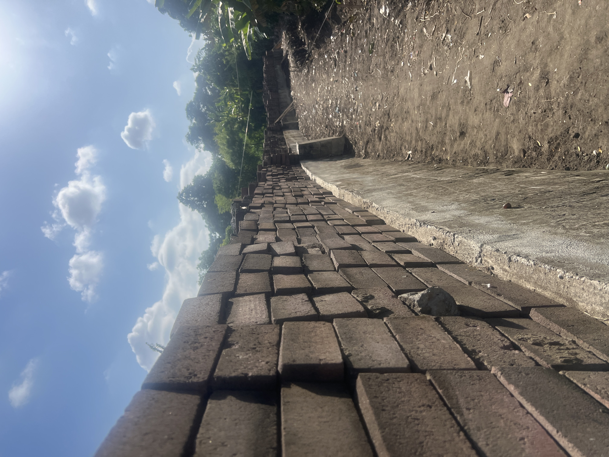 The image depicts a construction site in Denpasar, Indonesia, characterized by a long wall constructed of dark brown bricks. The bricks are stacked uniformly, forming a substantial barrier. Adjacent to the brick wall is a dirt path strewn with debris, suggesting ongoing or recent construction activity. The path leads towards a densely vegetated area in the background, with lush green trees and foliage visible under a bright, partly cloudy blue sky. The sun is shining, casting shadows and creating a slightly overexposed effect in the upper right portion of the frame. There are no people or animals visible in the scene. The overall impression is one of an outdoor construction area in a tropical climate.