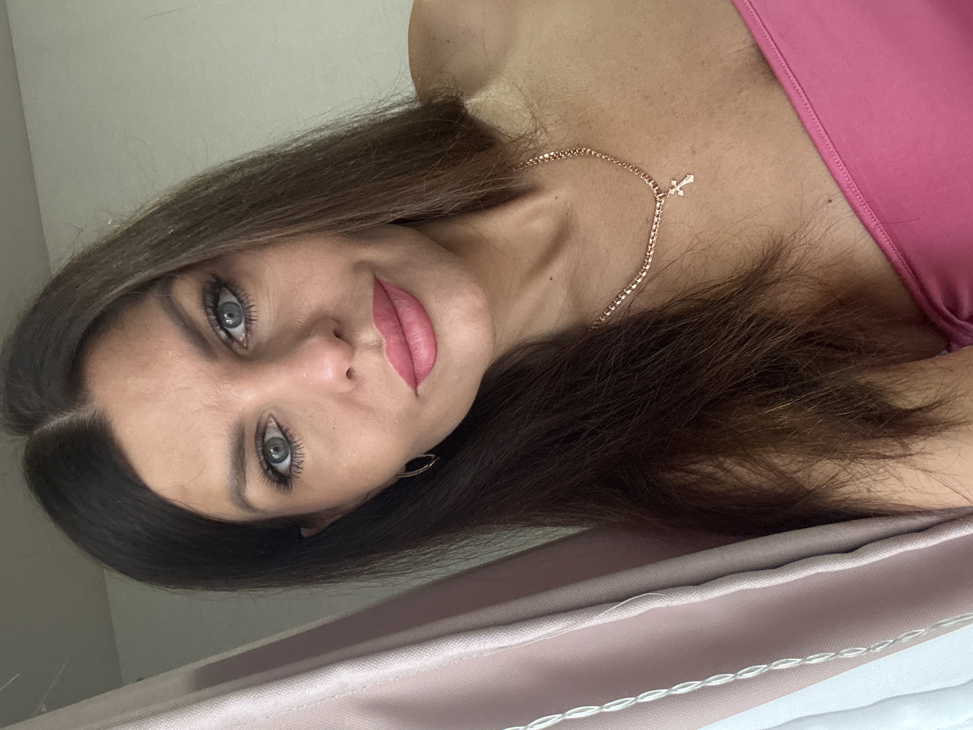 A woman with long brown hair and blue eyes is shown from the chest up in a selfie. She is wearing a pink strapless top and a rose gold necklace with a cross pendant. She is also wearing small gold hoop earrings. The background is a plain wall with a pink curtain on the left side of the frame. The image appears to have been taken indoors, possibly in a bedroom or living room. The woman is looking directly at the camera with a slight smile.