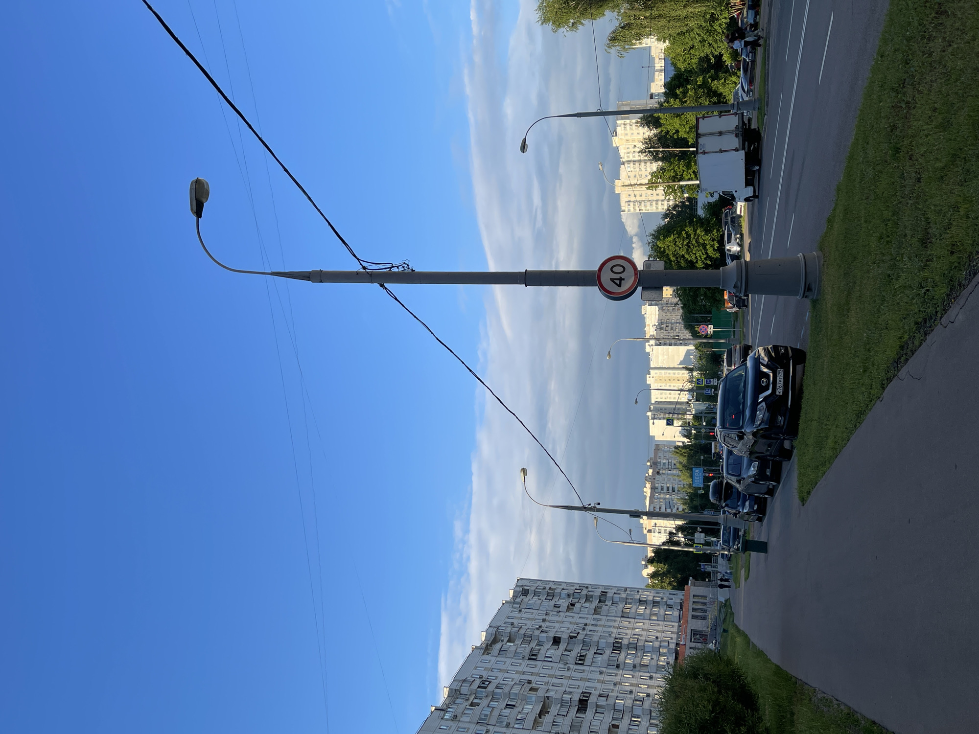 A street scene in Moscow, Russia, shows a wide sidewalk with a grassy verge on the left and a road with multiple lanes on the right. The sky is blue with some scattered clouds. A tall, gray lamppost with a curved arm holding a light fixture stands prominently in the foreground, accompanied by a speed limit sign indicating 40. Several parked cars line the grassy verge between the sidewalk and the road. In the background, multiple residential buildings of varying heights are visible, along with trees and other streetlights. The scene appears to be captured during daylight hours, likely late afternoon or early evening given the lighting.