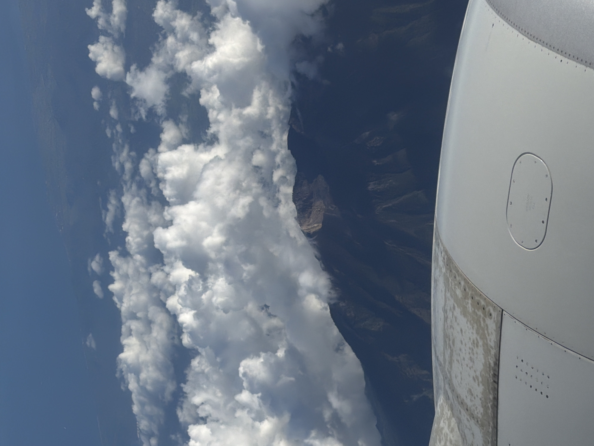 The image is taken from an airplane window. The right side shows a section of the airplane's wing, which is white. The exterior of the wing shows signs of wear. Visible outside the window is a landscape consisting of a blue sky on the left with fluffy white clouds, mountainous terrain partially shrouded in shadow, and possibly a distant volcanic peak visible between the clouds. The location is likely near Sukadana, Indonesia, as indicated in the instructions, considering the landscape's characteristics.
