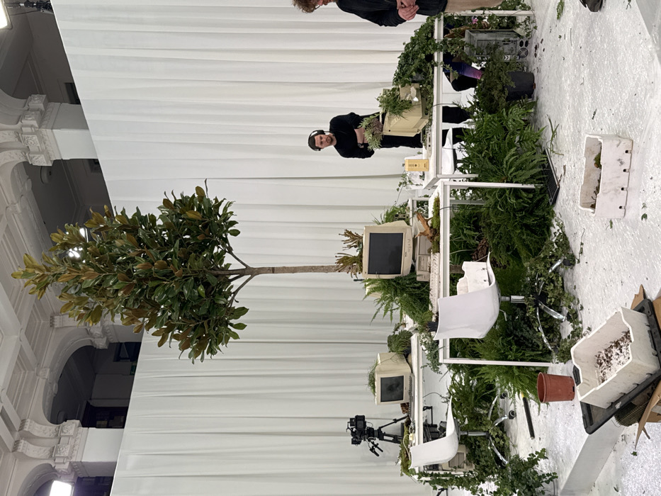 This image depicts a contemporary art installation within a large, high-ceilinged indoor event space or gallery in Milan, Italy. The background features white, floor-to-ceiling drapes that partially obscure ornate cream-colored architectural details, including arches and moldings. The floor is white, with scattered green plant debris.
Centrally, a tall, slender tree with dark green leaves stands amidst an arrangement of white rectangular desks. These desks are integrated with vintage beige computer equipment, including at least four CRT monitors, several keyboards, and desktop towers, all heavily overgrown with various green plants, ferns, and vines. Some plants appear to sprout directly from the technology casings. Two white modern office chairs with casters are positioned at two of these desks. A professional video camera on a tripod rests on the leftmost desk.
Two males are present in the scene. One, with dark hair and wearing a black long-sleeved shirt and over-ear headphones, stands behind a desk to the right of the central tree, with his arms crossed. The second male, with curly light brown hair, is partially visible on the far right. On the floor in the foreground, a brown plant pot and two white plastic trays containing plant matter are visible, with one tray resting on a cardboard box. An overhead rectangular studio light fixture is visible in the upper left corner.