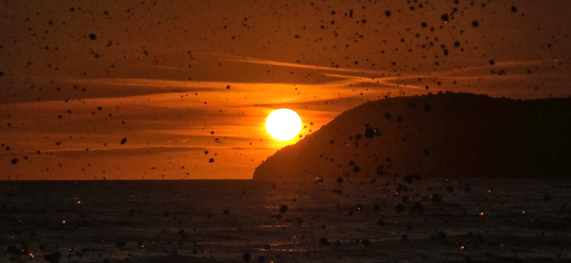 The image depicts a sunset over the Adriatic Sea from the city of Bar, Montenegro. A landmass, likely a coastal hill or mountain, occupies the right side of the frame. The sun, a bright white circle, is positioned near the peak of the landmass. The sky is orange, with subtle cloud formations. Water droplets, possibly on a lens or window, are visible as dark spots throughout the image. The sea reflects the orange hue of the sky, with faint glimmers of light. The overall scene appears dark and warm.