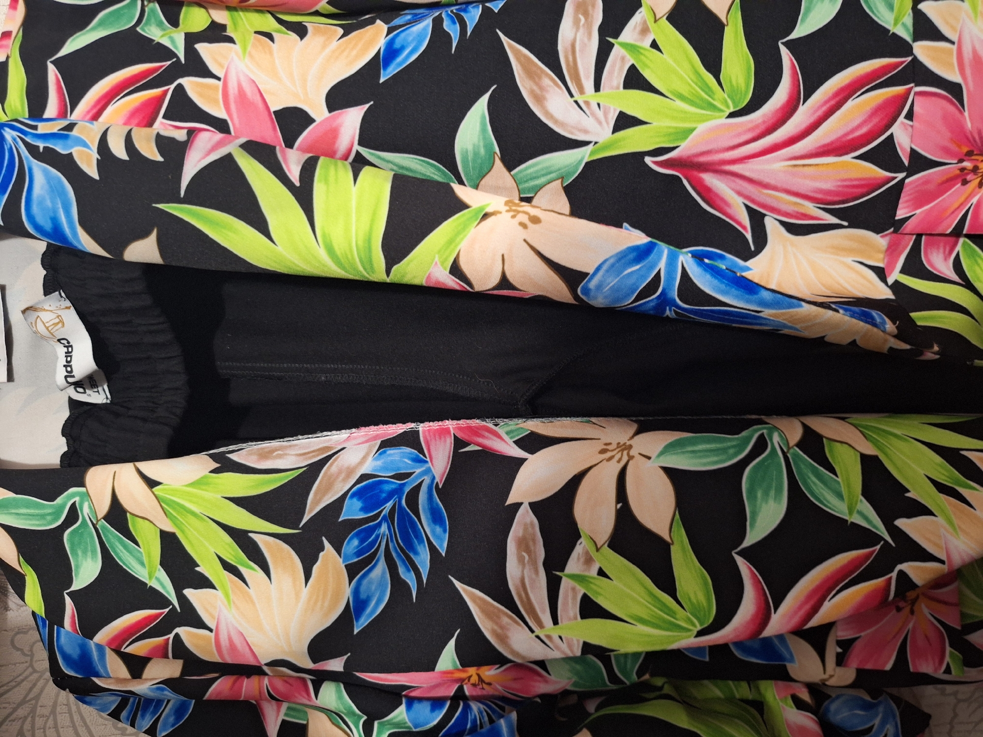 A floral print blazer in shades of black, green, pink, orange, and blue is draped over a black garment. A white tag with black text, partially obscured, is visible on the inside of the blazer's collar. The background appears to be a textured surface, possibly a wall with a light-colored wallpaper. The image is a close-up shot, focusing on the texture and patterns of the clothing. The scene is indoors, with no indication of an outdoor environment or any specific city location.