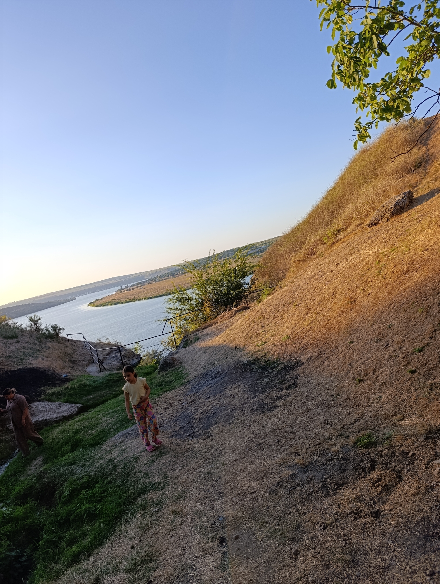 In Roghi, Moldova, a young girl in a yellow t-shirt and colorful pants stands on a dry, grassy slope facing a body of water. Behind her, a person in a brown outfit walks down a grassy incline towards the water's edge. The landscape features a wide river or lake with a distant, green shoreline dotted with trees and structures. The sky is clear and blue, with the warm light of what appears to be late afternoon or early morning illuminating the scene. A large, dry, grassy hill dominates the right side of the frame, with a few rocks and sparse vegetation. A tree branch with green leaves extends into the upper right corner of the image.