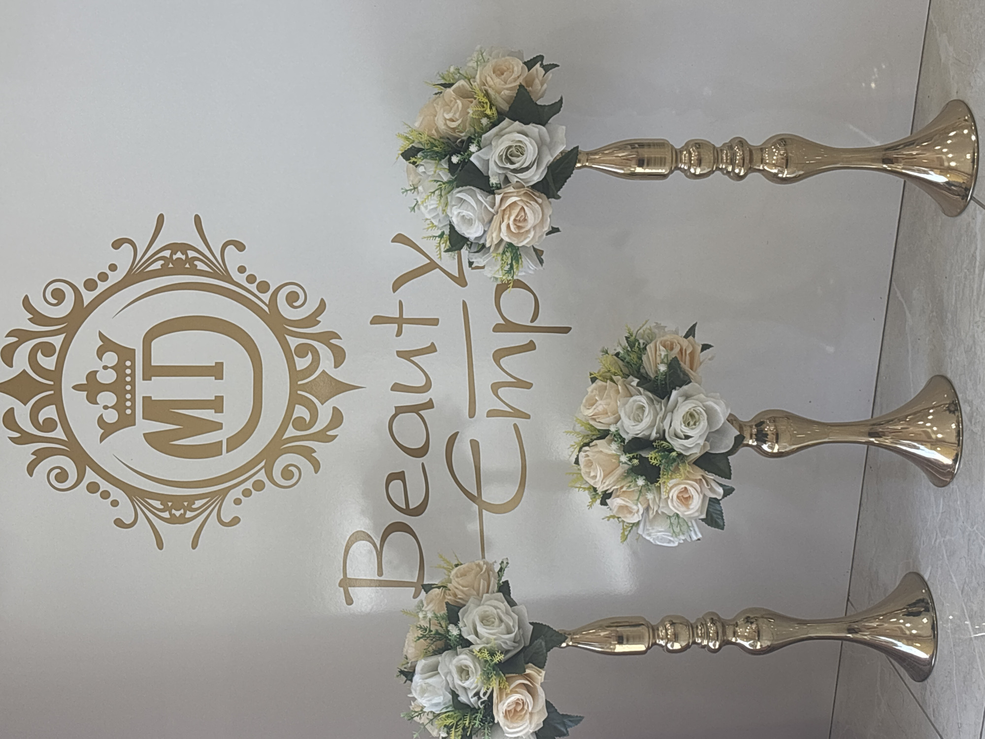 The image displays an interior setting, possibly a beauty salon, event space, or shop, located in Sofia, Bulgaria. The primary subjects are three floral arrangements on tall, golden, ornate stands, positioned against a light-colored, possibly white or off-white, wall. The floor appears to be a light-toned tile or polished surface.
The floral arrangements consist of artificial or silk flowers in soft hues of white, cream, and very light beige or blush, interspersed with dark green leaves and light green filler foliage. Each bouquet is dense and rounded. The golden stands are elegant, featuring a flared base, a slender, segmented body with decorative bumps and rings, and a cup-like top designed to hold the floral arrangements. They vary slightly in height, with one appearing taller than the others, and are arranged somewhat diagonally across the frame.
Prominently displayed on the wall behind the floral arrangements is golden decorative signage. To the left, there is an elaborate oval emblem, styled with a crown at the top and intricate scrollwork, enclosing the bold, stylized letters "MD". To the right of this emblem, the words "Beauty Emp" are written in a flowing, calligraphic golden font. The word "Emp" is incomplete, possibly standing for "Emporium" or "Empire," suggesting the name of the establishment. The overall aesthetic is one of elegance and sophistication, likely for a business focused on beauty or events. There are no people visible, and no specific activity is taking place; the image captures a decorative setup.
