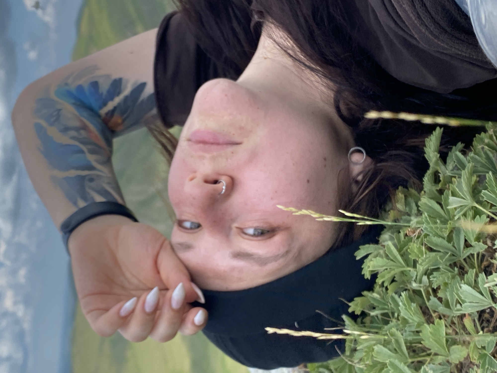 A close-up, low-angle shot shows a person lying down in a grassy, natural environment. The person is wearing a black beanie and a dark brown garment. Their right arm, adorned with a blue, feather-like tattoo, is bent, with their hand resting on their forehead. They have a nose ring and an earring. In the foreground, green foliage with thin, light-colored stalks is visible. The background depicts rolling hills under a cloudy sky, suggesting an outdoor setting in a rural area, potentially near the village of Abzanovo, Russia. The lighting is natural, likely daylight, and the focus is sharp on the person and the immediate surroundings.