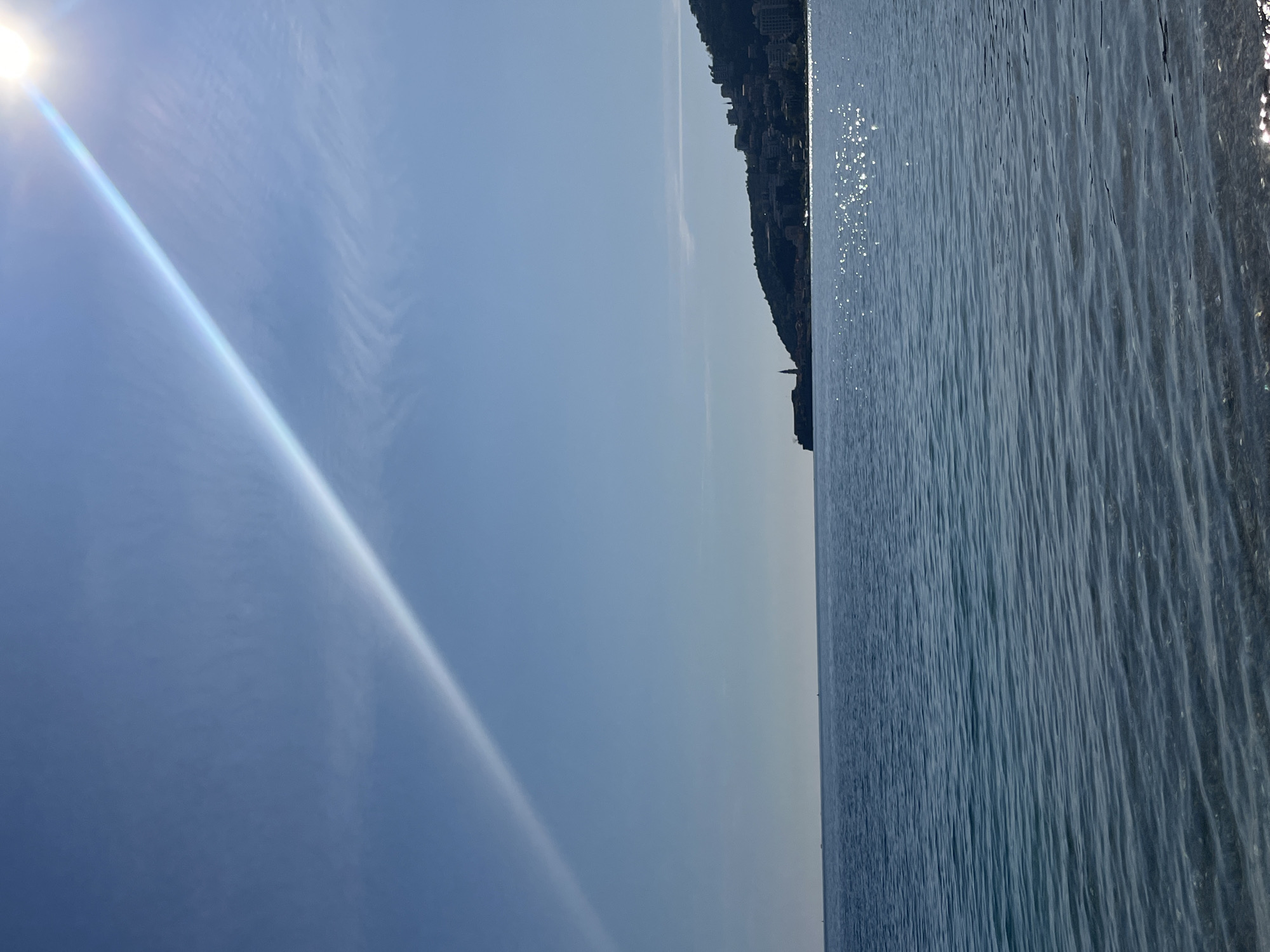 The image captures a daytime scene in Budva, Montenegro. The sea occupies a significant portion of the view, its surface rippling with small waves reflecting sunlight. Along the horizon, a heavily built-up hillside is visible, suggesting the urban character of Budva. The sky above is light blue, with a bright sun causing a glare. The angle of the shot suggests it was taken from a low position, potentially from the water or near the shoreline. There are no visible people.