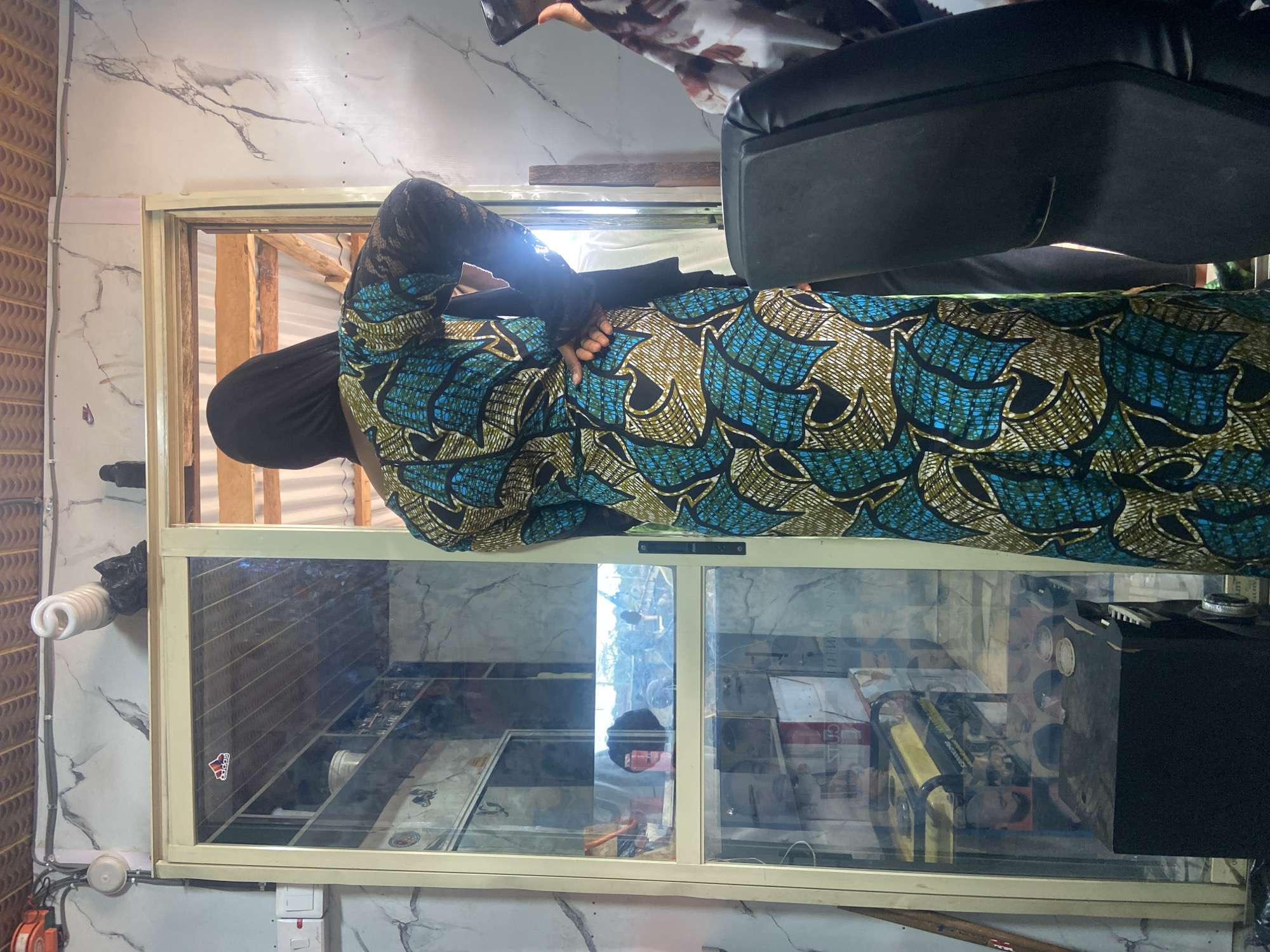 An individual wearing a traditional, patterned dress and a head covering is seen from behind, standing near a glass-fronted display case. The case appears to contain various objects, including packaged goods and what seems to be an electronic device. The surrounding environment consists of a building interior with patterned walls and a view through an open section that reveals wooden beams and framing. The location is Okengwe - Obehira, Nigeria. A black chair with a black cushion is visible. Electrical wiring and fixtures are visible. An Adidas sticker is placed on the left side of the glass-fronted display case.