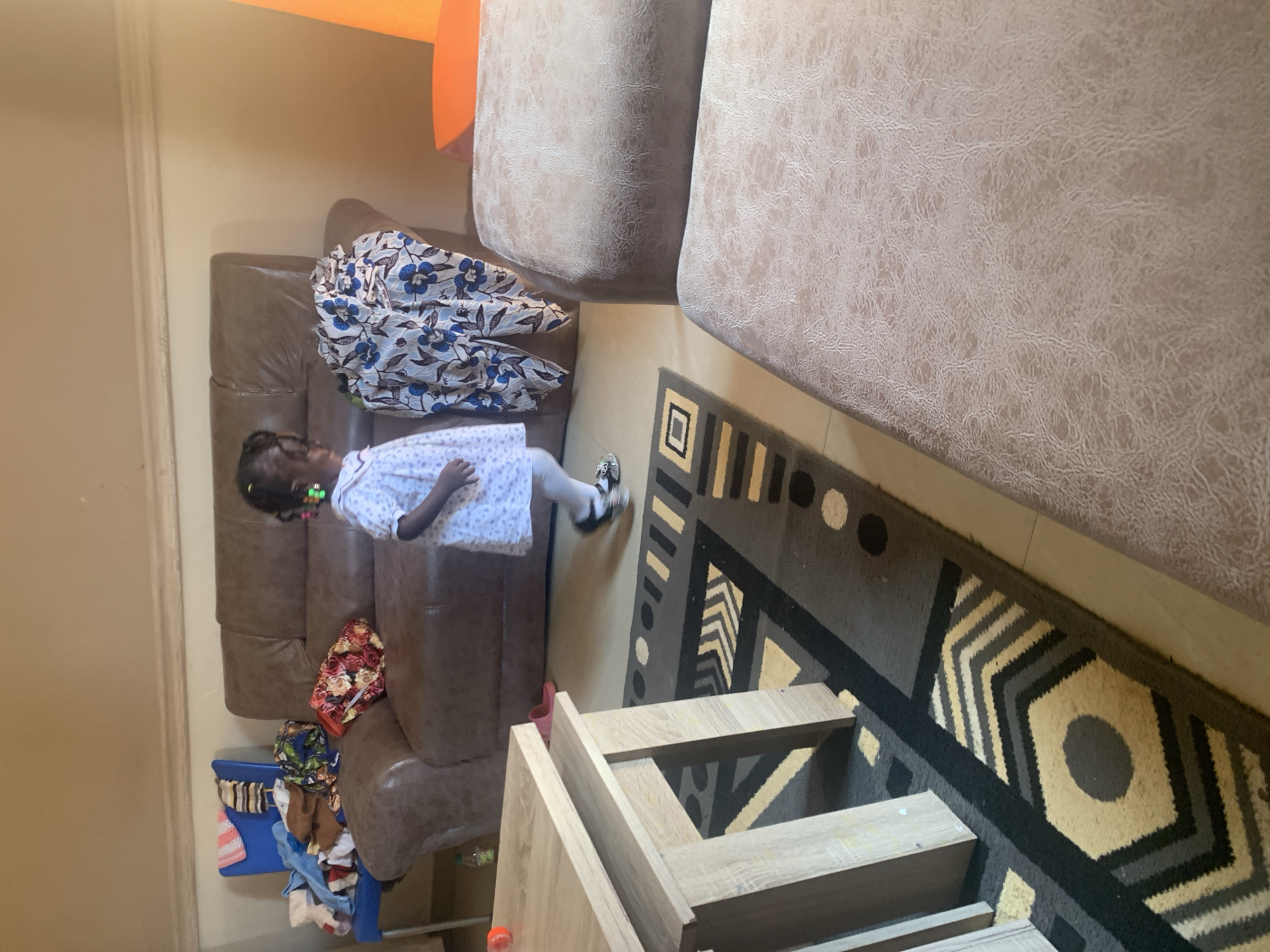 The image depicts an indoor scene, likely a living room, in Gura Top, Nigeria. A young child, appearing to be a girl, is walking towards the right side of the frame. She is wearing a white dress with a patterned print and white tights. Her hair is styled in braids and she is wearing green earrings.
In the background, a large brown leather sofa is visible, with various items of clothing and fabric placed on it. To the left of the sofa, a blue chair holds more clothing items. A patterned rug with geometric designs in black, grey, yellow, and white is on the floor. A light-colored wooden coffee table is positioned on the left side of the rug, with an orange object on its surface. Two light-colored cushioned ottomans are in the foreground, to the right of the coffee table. The walls appear to be a light neutral color, with molding at the top.