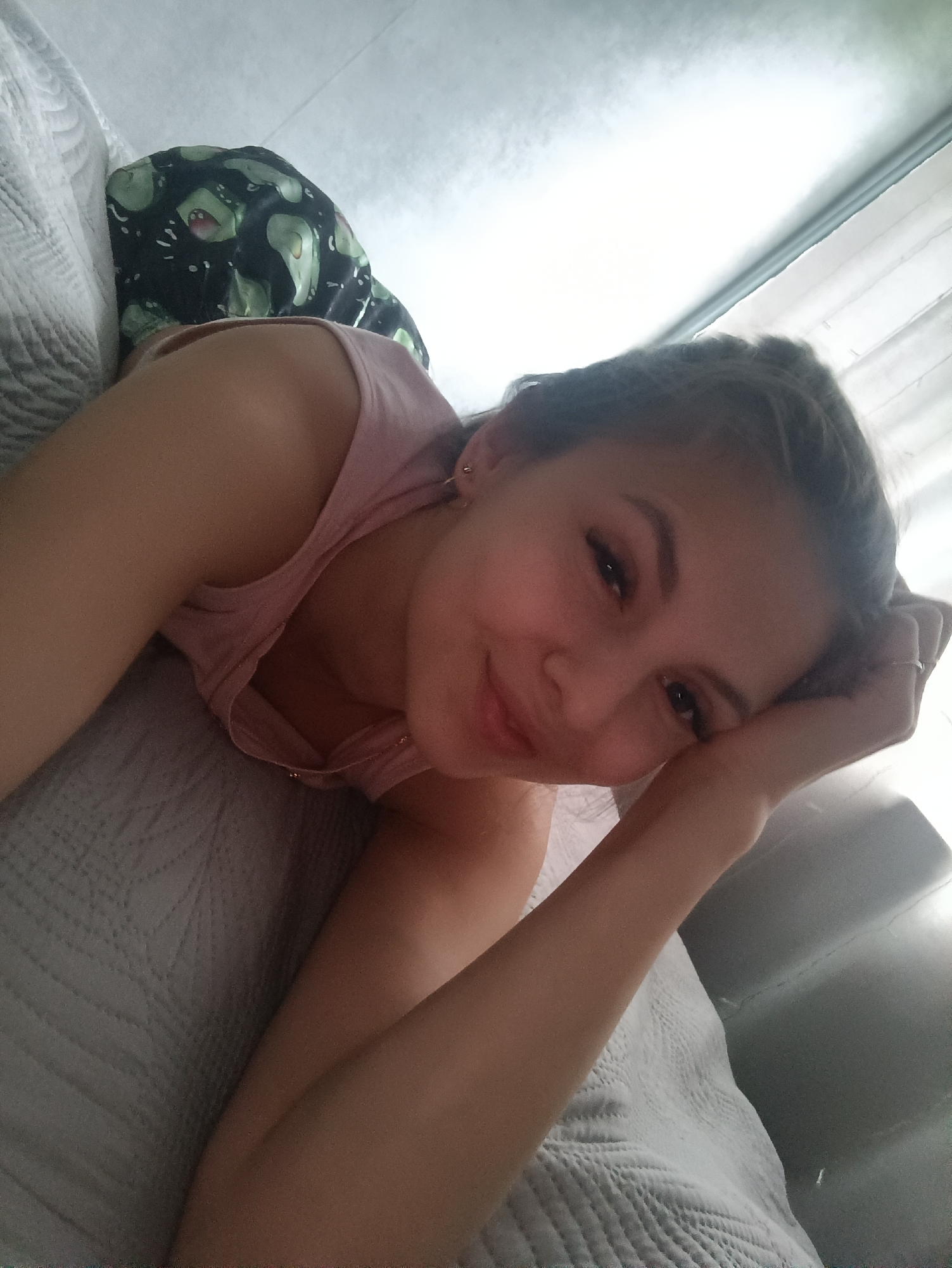 A woman lies on a bed with a gray quilt. She wears a light pink tank top. A black and green patterned garment rests on her back. Her head rests on her hand, and she smiles at the camera. A window is visible in the background. The location is unknown.