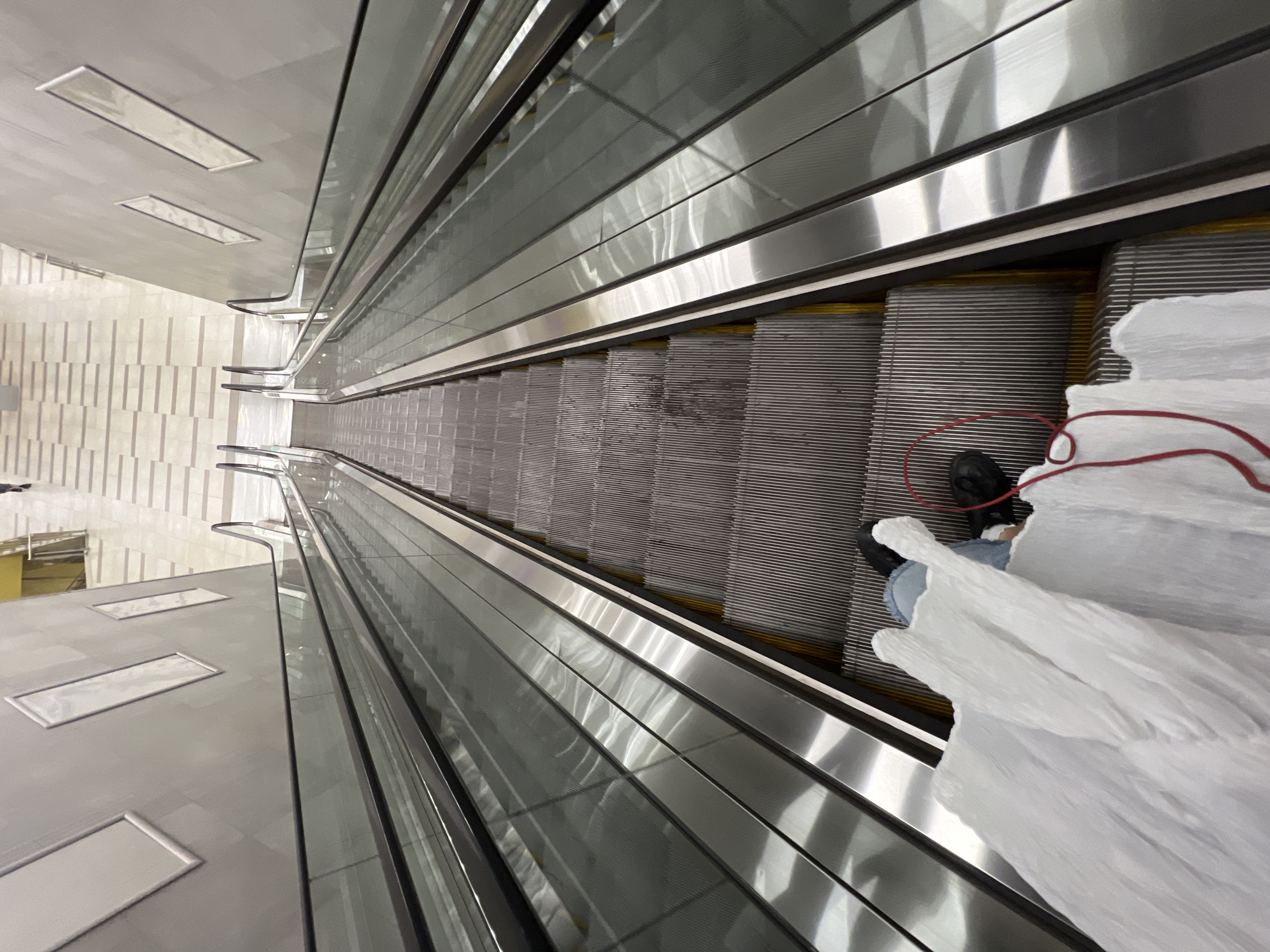The image shows a downward-facing view of two escalators. The escalator on the right is more visible and appears to be in motion, as indicated by the blurred steps and the reflective surfaces. The escalator on the left is partially obscured by a large white object in the foreground. A person's legs and feet are visible on the left escalator, wearing blue jeans and black shoes. A red cord is also visible, coiled on the escalator steps near the person. The walls of the escalator shaft are lined with a gray material and feature several rectangular decorative panels. The floor at the bottom of the escalators is tiled in a light beige and white striped pattern. The overall impression is that of a modern, indoor public space, possibly a shopping mall or airport, and it is unlikely to be specifically in Sofia, Bulgaria, as there are no distinguishing features suggesting that location.