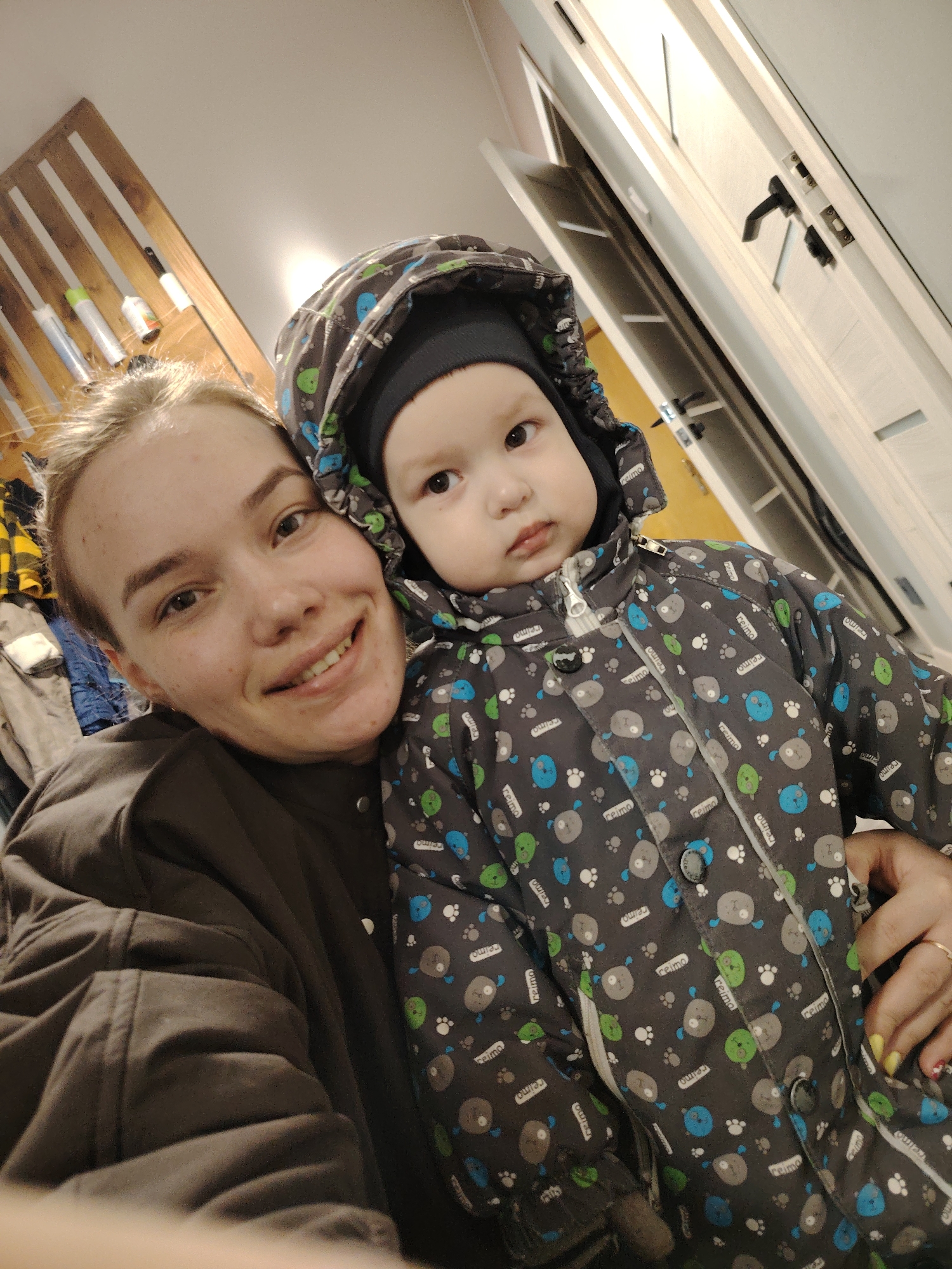 Here is a forensic description of the image:
**Scene:** An indoor selfie taken at an upward angle, likely in a home. There's a wall with wooden slats and what appear to be aerosol cans or similar items on the wall in the background. A white door is also visible.
**People:** A woman is holding a small child, possibly an infant or toddler. The woman is fair-skinned and has her hair pulled back. She is wearing a dark brown or black jacket. The child is bundled in a gray winter suit with a hood, which has a pattern of stylized dogs or bears and the brand "Reima" printed on it. The child is wearing a black cap under the hood.
**Subjects:** The woman is smiling and looking directly at the camera. The child has a more neutral or slightly serious expression.
**Objects:** A wooden wall decoration, aerosol cans, a door, the woman's jacket, the child's winter suit, and the child's hat.
**Actions:** The woman is taking a selfie while holding the child close.
**Interactions:** The woman is holding the child, and they are both looking towards the camera.
**Environment:** The scene appears to be in a residential setting, possibly an entryway or hallway.