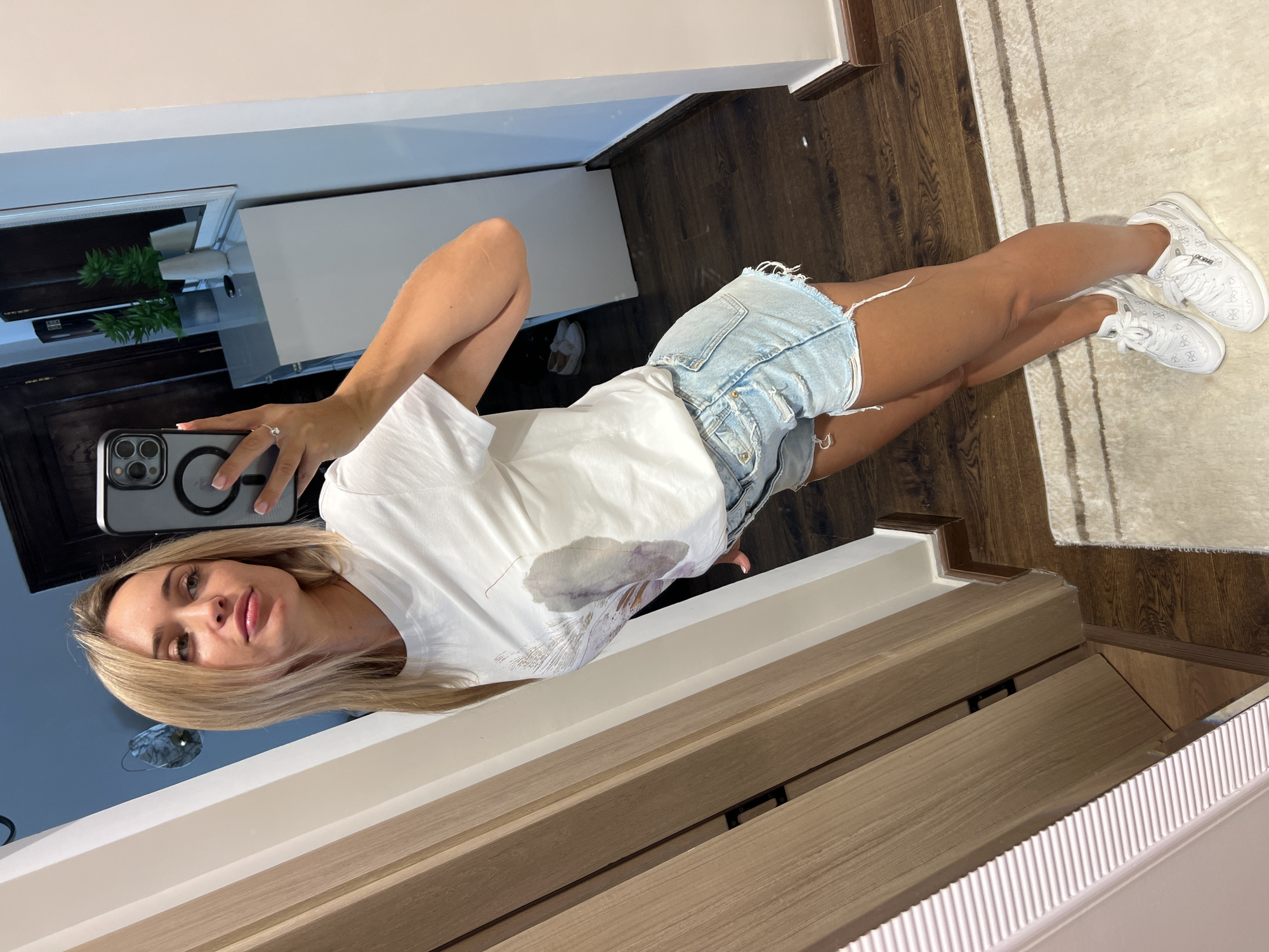 A blonde woman is taking a mirror selfie in what appears to be a hallway or dressing area. She is wearing a white t-shirt with a graphic print, light blue denim shorts with distressed detailing, and white sneakers. She is holding a smartphone in her right hand, which is visible in the mirror. Her left arm is resting on her hip. The background shows a wooden door on the left, a white cabinet with plants on the right, and a dark wooden floor with a light-colored rug. The scene is well-lit, suggesting an indoor environment. The presence of a mirror indicates this is likely a personal space such as a home or hotel room. The overall atmosphere is casual and relaxed. The location is not identifiable as Sharm El Sheikh, Egypt.