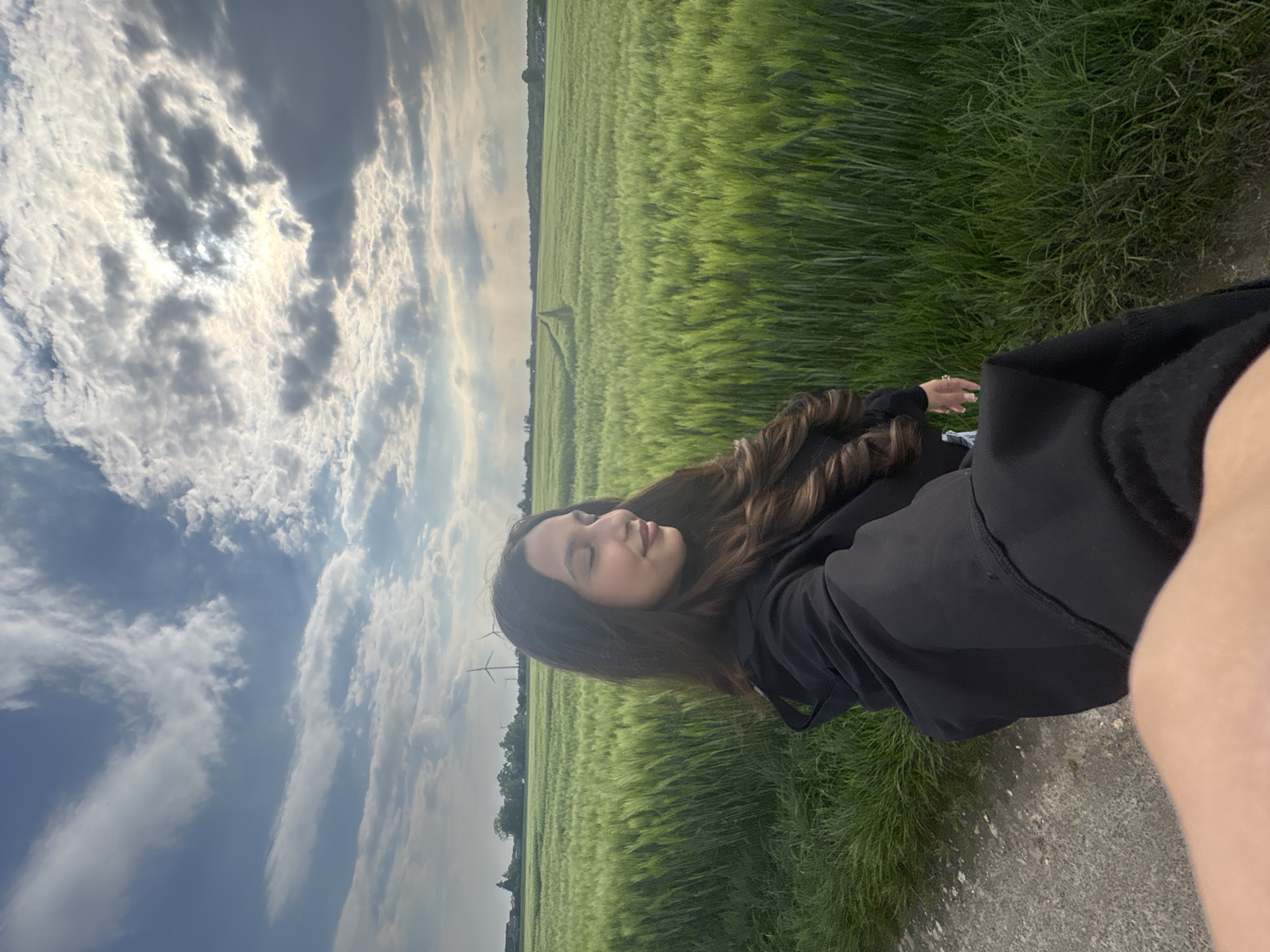 A medium shot shows a young woman posing for a selfie in a rural area near Wegberg, Germany. She is positioned on a narrow path or road bordering a vast field of green grain or grass. She is wearing a black sweatshirt, and her long, dark, wavy hair is partially visible. The field stretches out towards the horizon, partially obscured by a cloudy sky. In the distance, some trees and structures are visible. The sky is filled with a mix of white and dark clouds, with the sun peeking through in some areas. The angle of the shot suggests that the camera is held at an odd tilt, making the horizon appear diagonal.