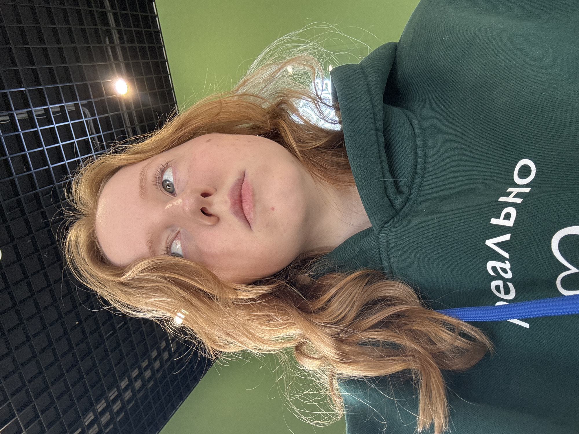 A close-up, low-angle photograph features a young person with fair skin and long, wavy auburn hair. They are wearing a dark green hooded sweatshirt with Cyrillic lettering visible. A blue lanyard or drawstring is partially visible on the sweatshirt. The background includes a black grid-patterned ceiling and a green wall. The photo appears to be taken indoors under artificial lighting. The location is uncertain as there is nothing in the image to suggest that this picture was taken in the city of Chaikovsky, Russia.