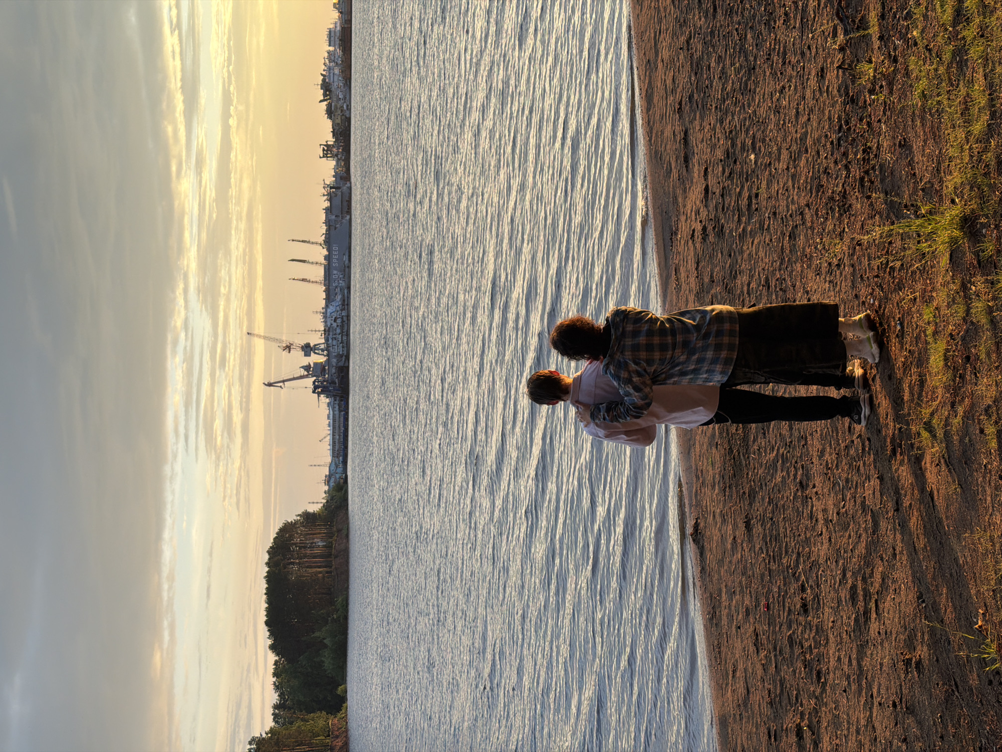 Two individuals, a male and a female, stand on a sandy embankment facing a body of water. The male, on the left, is wearing a light pink collared shirt and dark pants, with white sneakers. The female, on the right, is wearing a plaid shirt, a dark skirt, and light-colored shoes. Her right arm is around the male's shoulders. The water is calm, with small ripples reflecting the sky. In the background, across the water, is a tree-lined shore and a distant industrial complex with cranes and ships. The sky is a mix of clouds and golden sunlight, suggesting it is either sunrise or sunset. The scene appears to be at a waterfront location in the city of Chaikovsky, Russia, possibly near the Kama River, given the industrial activity and natural landscape.