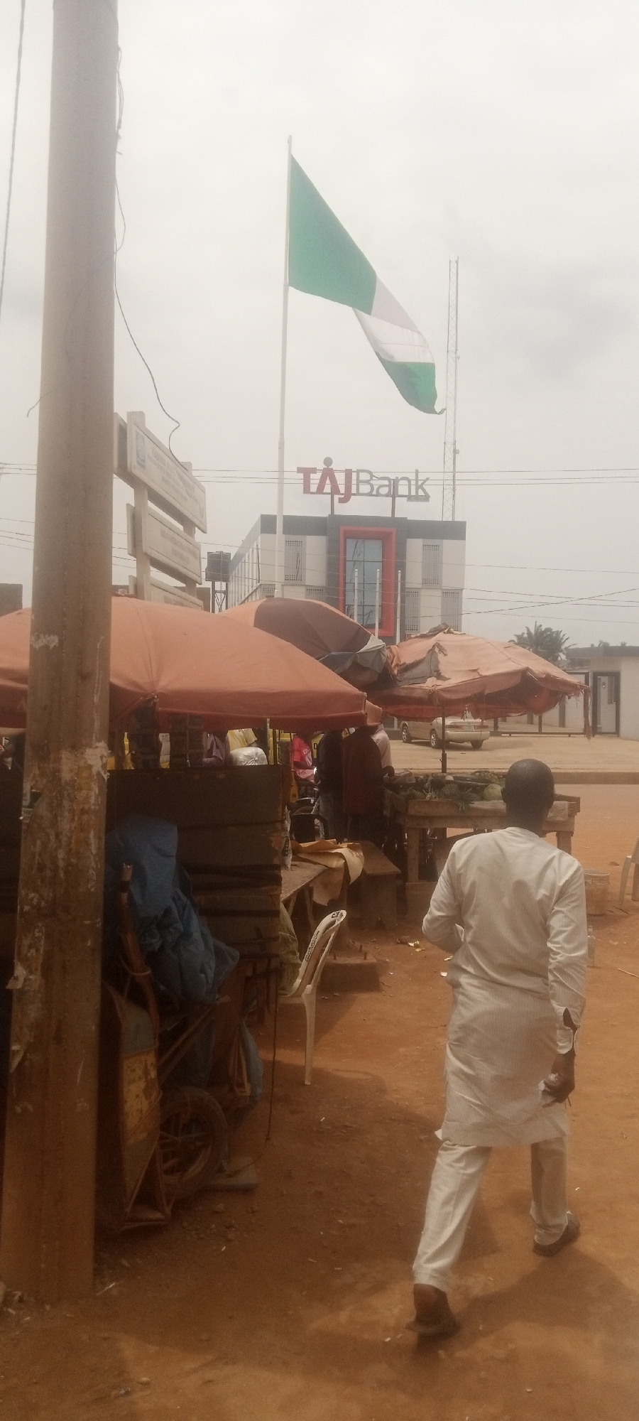 The image shows a street scene in Sabon Tasha, Nigeria. A man wearing a white shirt and pants walks away from the camera in the foreground. Behind him, there are market stalls covered with brown umbrellas, and a few people are standing around the stalls. In the background, a Nigerian flag is flying in front of a "TAJ Bank" building. A utility pole is on the left side of the image. The ground is reddish-brown.