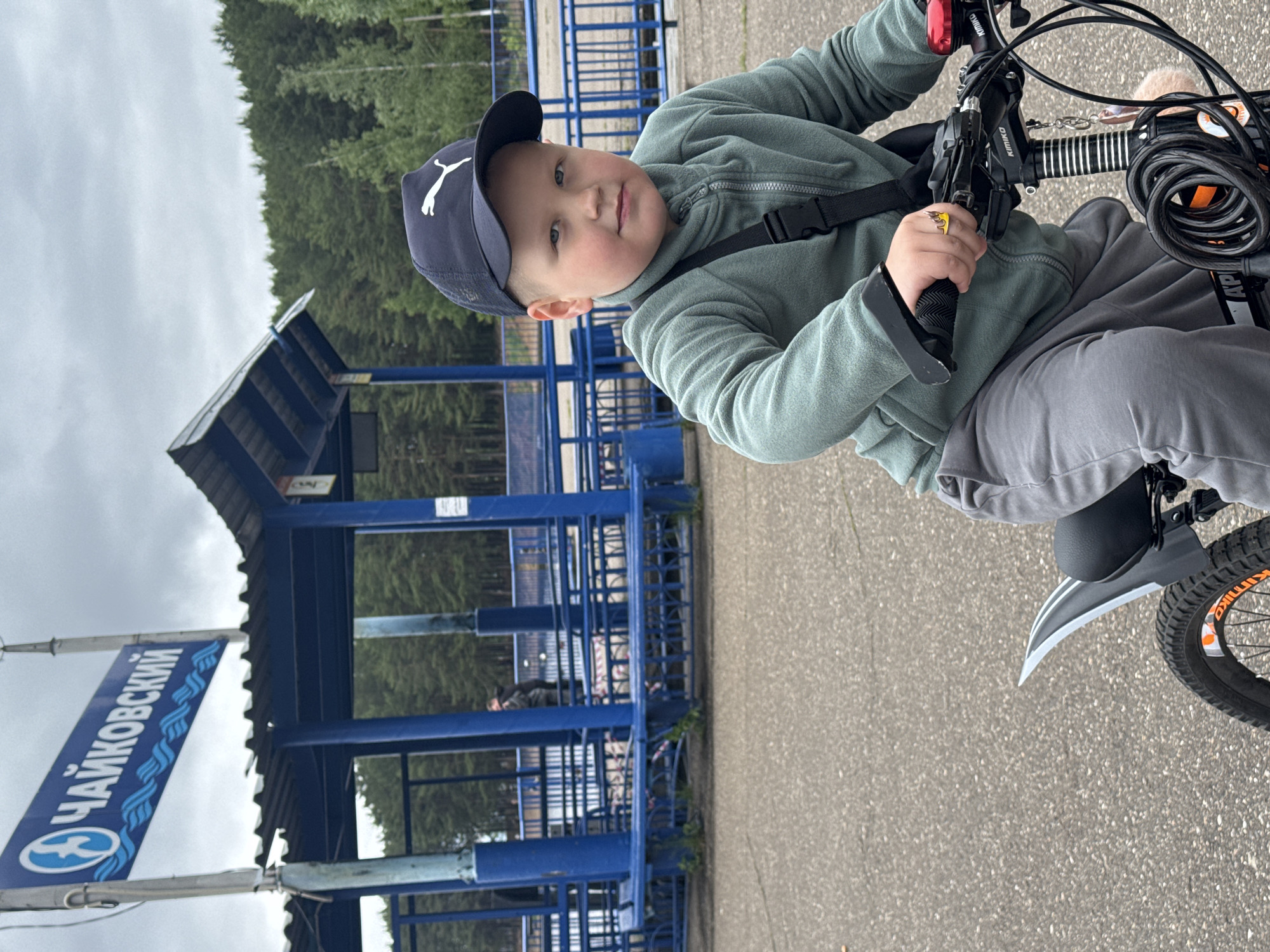 A young boy, approximately 6-8 years old, is seated on a black bicycle, positioned in the foreground and center-right of the frame, looking directly at the camera. He wears a dark blue baseball cap with a white Puma logo, a light green fleece zip-up jacket, and grey sweatpants. A black cross-body bag is slung over his left shoulder, and a yellow and orange ring adorns his right hand. The bicycle's handlebars show "KIMIKO" on the stem, a red bell on the right, and a black coiled cable lock attached centrally.
The scene is set in Chaikovsky, Russia, as indicated by a blue sign on the left side of the midground, which displays "ЧАЙКОВСКИЙ" in white Cyrillic letters above a stylized blue circular logo featuring a figure or bird over wavy lines. Behind the boy, a prominent blue-painted metal structure spans the midground horizontally. This structure consists of multiple vertical pillars and horizontal railings, forming what appears to be a covered walkway or a series of public gates/shelters. Two indistinct adult figures are visible behind this blue structure in the distance. The background features a dense forest under an overcast sky. The ground is paved asphalt.