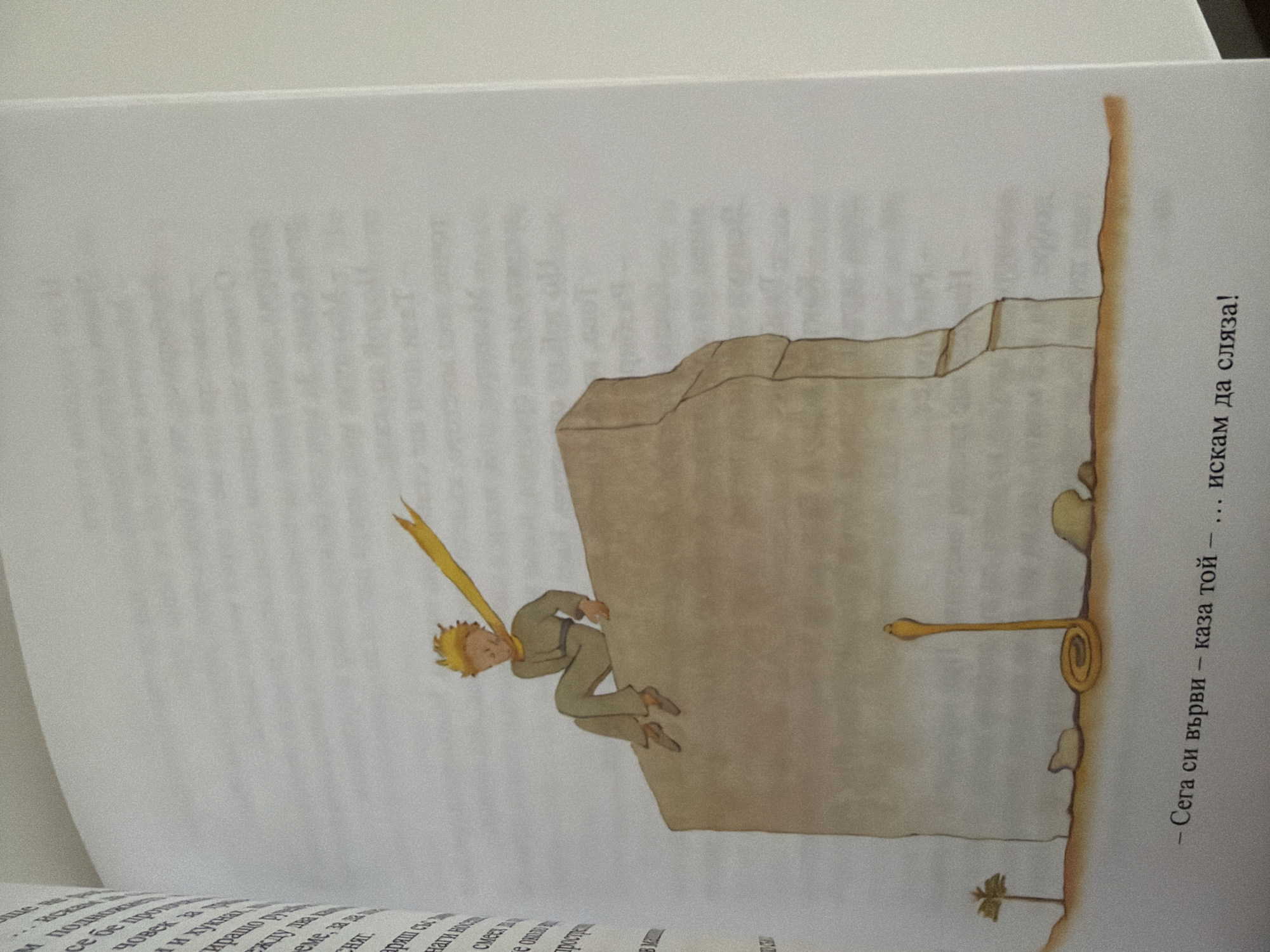 The image shows an illustration from a book. A young boy with blond hair and a yellow scarf is sitting on the edge of a large rock. Below the rock, a yellow snake is coiled. The text at the bottom of the image reads, "— Сега си върви — каза той — ... искам да сляза!". This is a scene from "The Little Prince." There is no information in the image that suggests a location in Pazardzhik, Bulgaria.