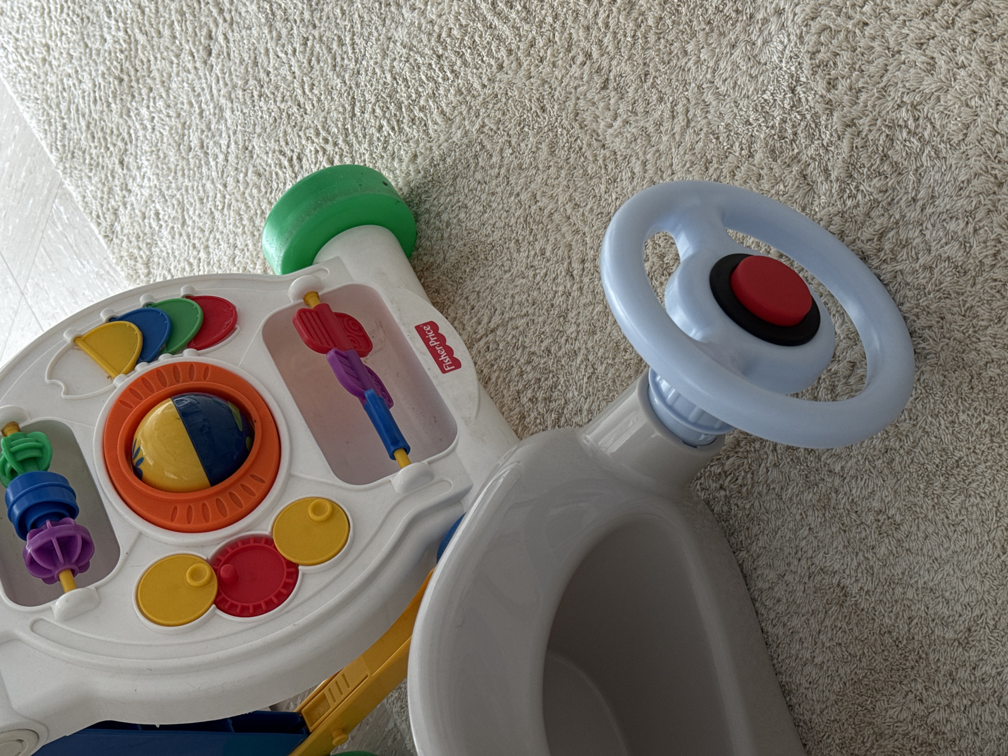 This is a close-up, eye-level, indoor, medium shot of a partially visible Fisher-Price activity walker toy. The toy is white with colorful accents, including yellow, red, blue, and green components. The foreground is dominated by a light gray, oval-shaped seat area of the walker and a light blue steering wheel with a red button in the center. The background consists of a beige, textured carpet. The scene appears to be a domestic environment. There are no people present in the image. The lighting is neutral and even, suggesting ambient indoor light. The toy is not in motion. There is no indication of the specific location within Siegburg, Germany.