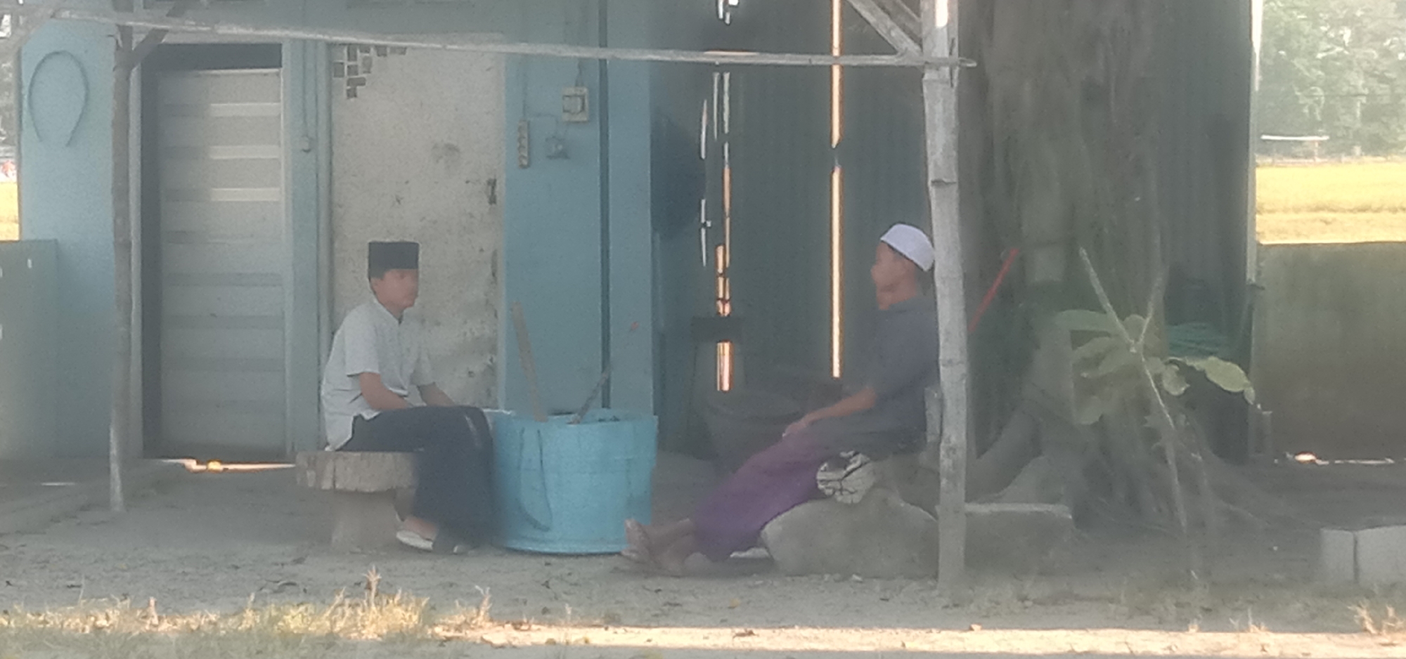 Two individuals are seated outdoors. The person on the left, appearing to be a young male wearing a black skullcap and a light-colored collared shirt with dark pants, is seated on a rough-hewn bench made of wood. To his right, a blue barrel-like object is visible. The person on the right, an adult male wearing a white skullcap and a dark shirt with purple pants, is seated on a large stone or concrete structure. They are positioned within an open-sided structure with visible wooden supports and a tin roof. Behind the structure, a pale blue wall and a doorway are discernible. Further in the background, a field of yellowing crops can be observed, suggesting a rural or agricultural setting. The overall scene is casual and appears to be in a village or small community.