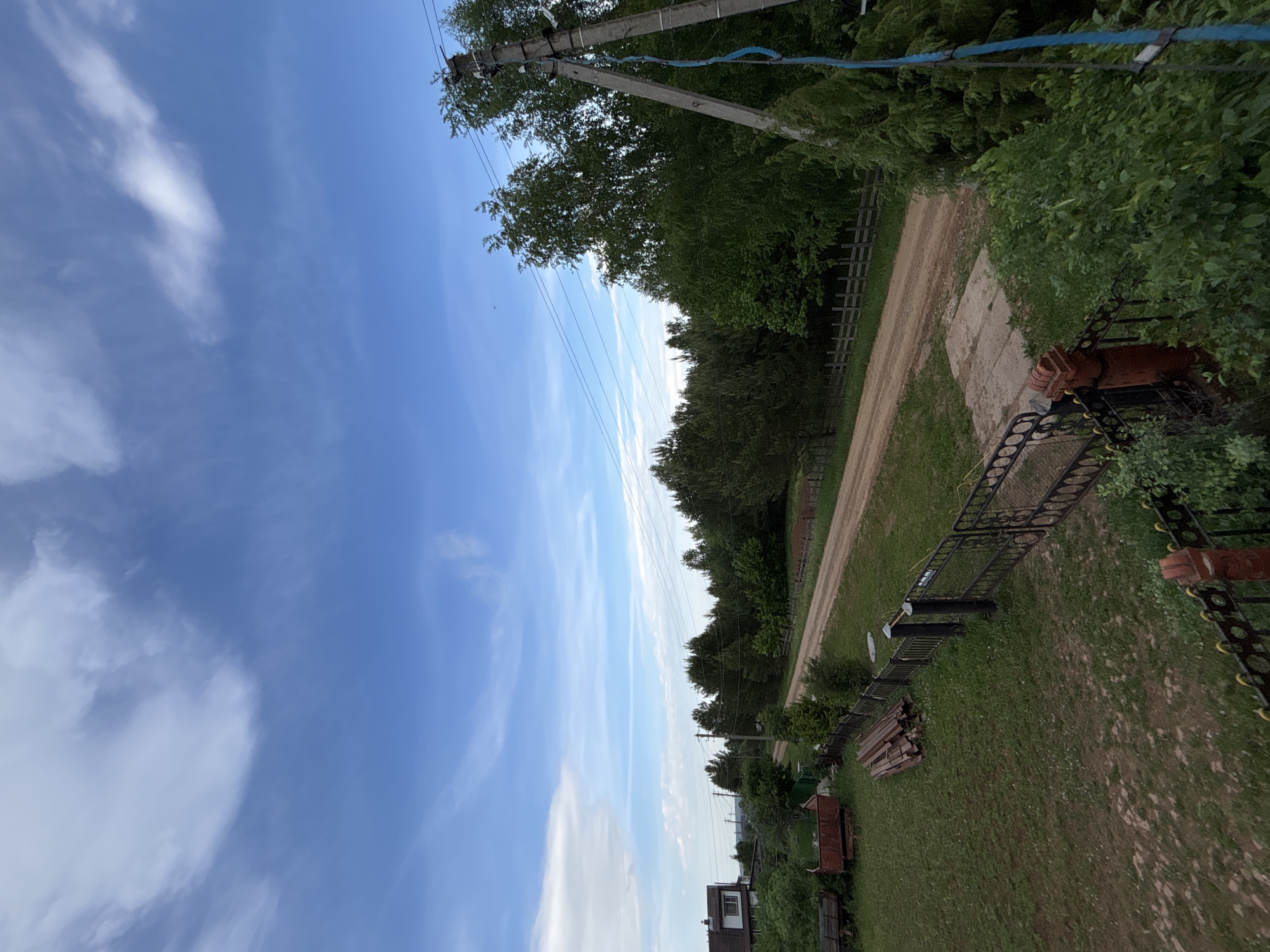 The image depicts a rural scene in Большой Букор, Russia. A dirt road is flanked by trees and vegetation. Power lines run along the road. A fence and other objects, possibly farm equipment, are present in the foreground. A house is visible in the distance. The sky is blue with clouds.