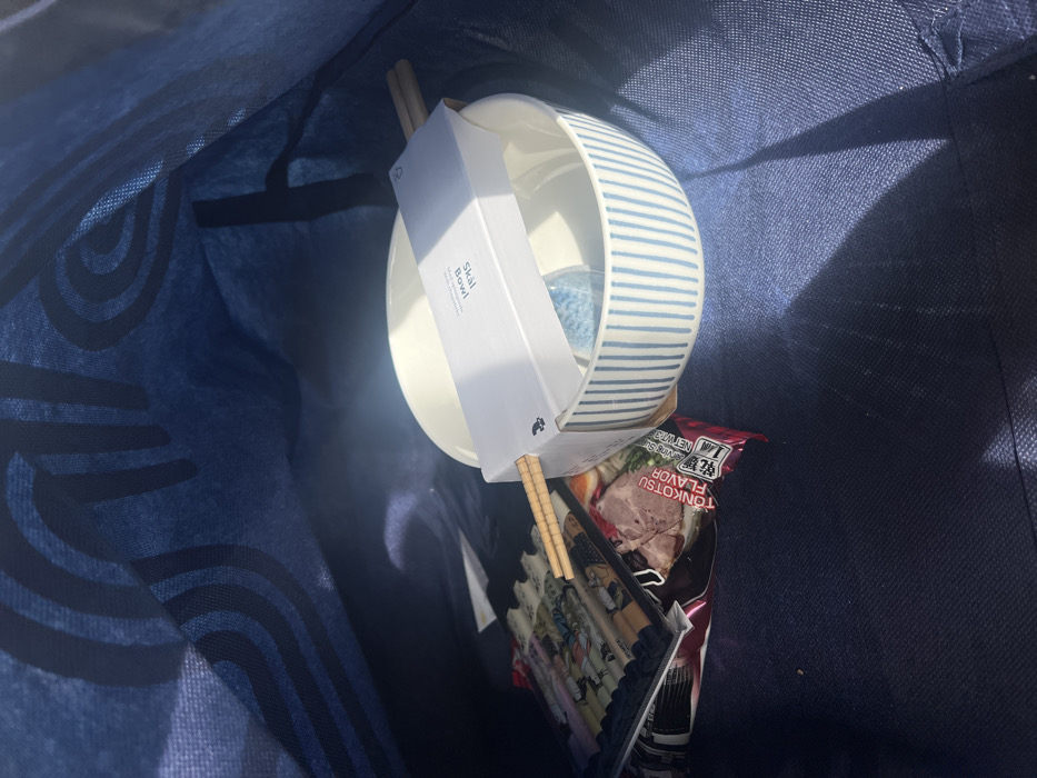 The image displays the interior of a dark blue fabric bag, set within the Hof Ashkelon Regional Council, Israel. The bag's inner lining features a repeating pattern of darker blue curved lines. Strong, direct light from the upper right illuminates the contents, casting shadows to the left.
Within the bag, two ceramic bowls are stacked centrally. The upper bowl is plain white. The lower bowl is white with vertical light blue stripes. A white cardboard sleeve labeled "Skal Bowl" is positioned horizontally across the top of the stacked bowls, securing a pair of light-colored wooden chopsticks. A small, light blue spherical object is visible inside the bottom bowl.
To the left of the bowls, a red and white packet marked "Tonkotsu Flavor" (indicating instant ramen) is partially visible. Below this packet is a stack of several flat, rectangular items displaying decorative patterns. One of the chopsticks rests across the top of this stack of items. No people are present.