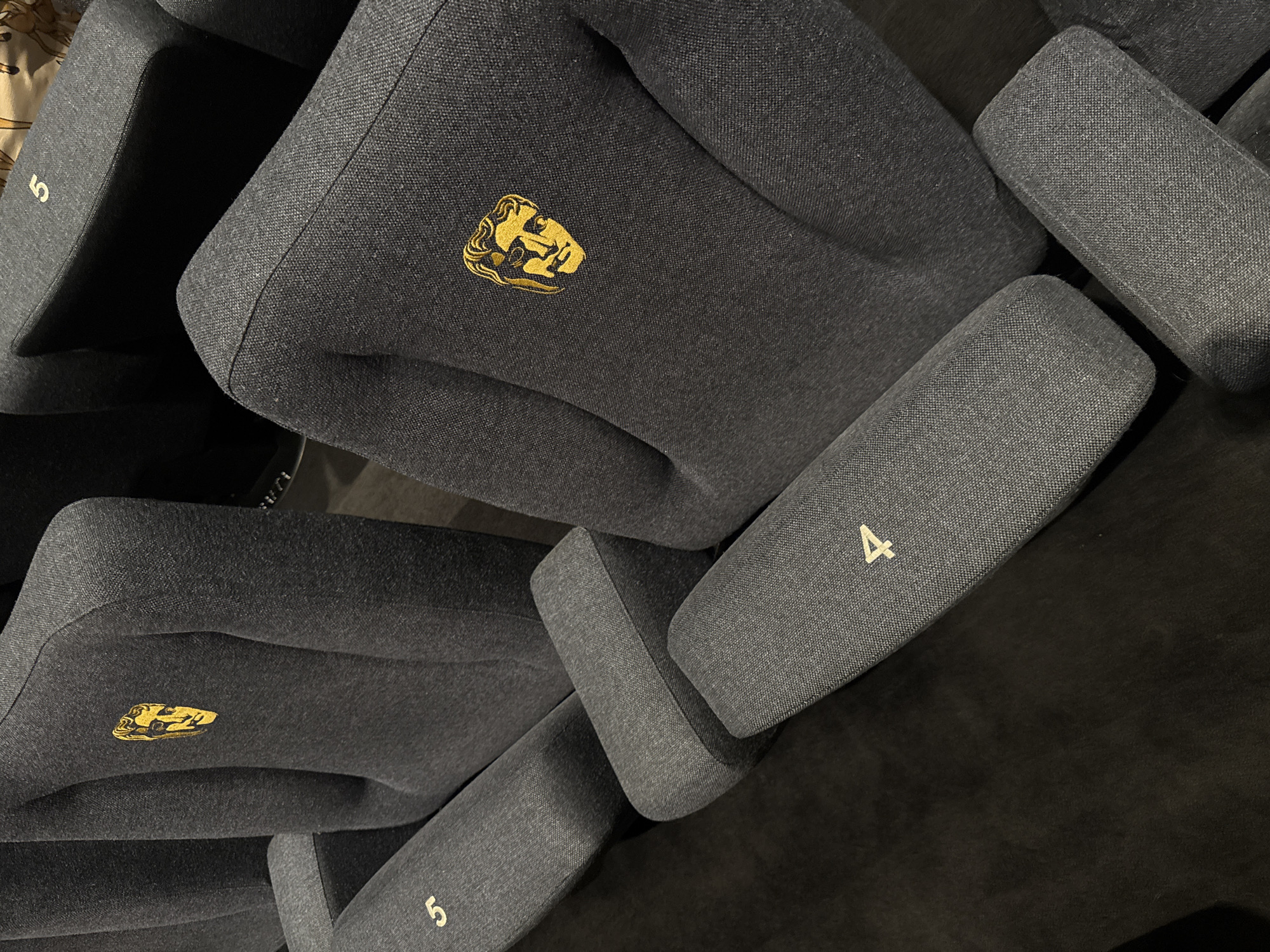 This image captures a close-up, slightly angled view of several dark gray upholstered seats in an indoor setting, likely an auditorium or cinema in London. The seats feature a textured fabric, possibly a wool or linen blend, and are arranged in rows, though only parts of a few seats are visible.
A distinctive gold-embroidered logo, depicting the stylized mask of Tragedy, is prominent on the backrest of one central seat. This emblem is identifiable as the British Academy of Film and Television Arts (BAFTA) logo, strongly indicating the location is a BAFTA venue.
Gold numbers are visible on the armrests of some seats. The number "4" is clearly seen on the armrest of a seat to the right, and two instances of the number "5" are visible on armrests to the left and bottom-left, suggesting a seating arrangement with designated numbers. The seats appear empty, indicating no specific activity is currently taking place. The lighting is artificial and somewhat dim, typical of an indoor entertainment or conference space. The focus is on the design and branding of the seating, highlighting the connection to BAFTA.