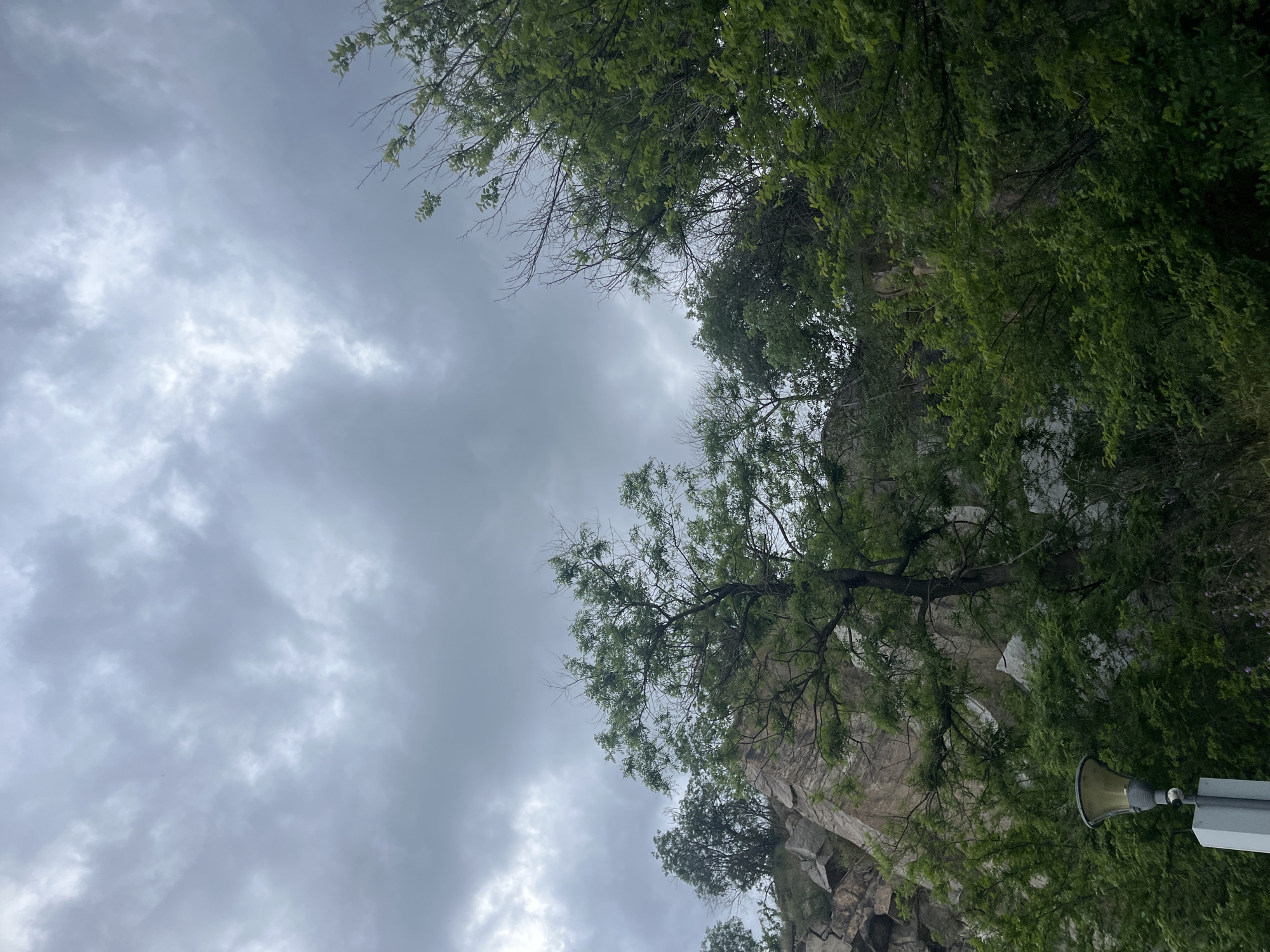 The image shows a cloudy sky and a rock face with trees, taken from an upward-looking perspective. At the bottom right corner is a loudspeaker mounted on a pole. The location appears to be Nebet Tepe in Plovdiv, Bulgaria.
