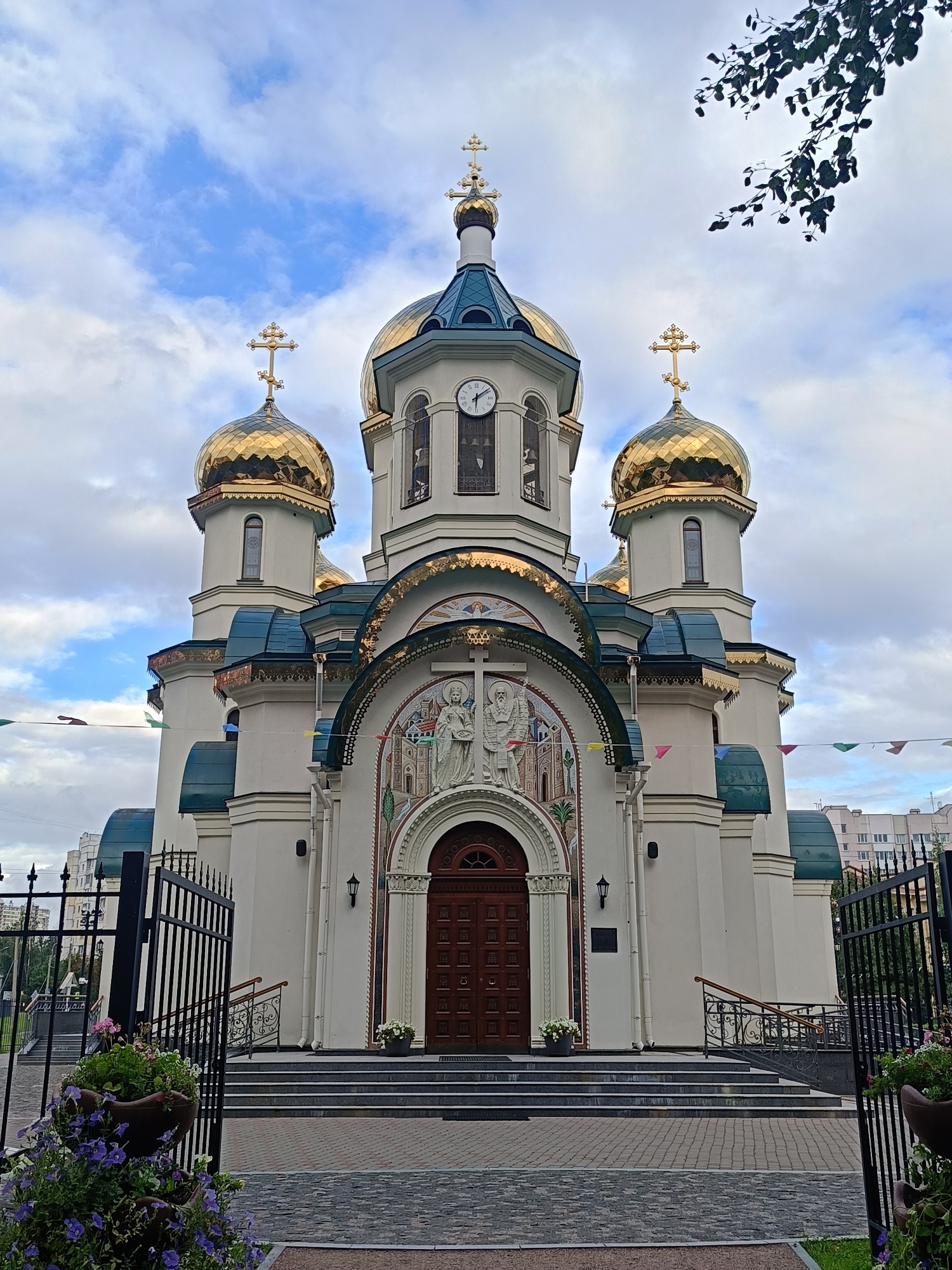 This is a low-angle, full-shot photograph of the Saint Petersburg Mosque, located in Saint Petersburg, Russia. The mosque is a large, multi-domed structure with a white facade and gold domes. The main dome is centrally located, with smaller domes on either side. The mosque has a large wooden door at the entrance, with intricate mosaic work above it. There are also two minarets flanking the main entrance. The mosque is surrounded by a black wrought-iron fence, and there are steps leading up to the entrance. Flower pots with purple and pink flowers are placed in front of the fence. The sky is cloudy, with patches of blue visible. The mosque is a prominent landmark in the city, and it is a popular tourist destination.