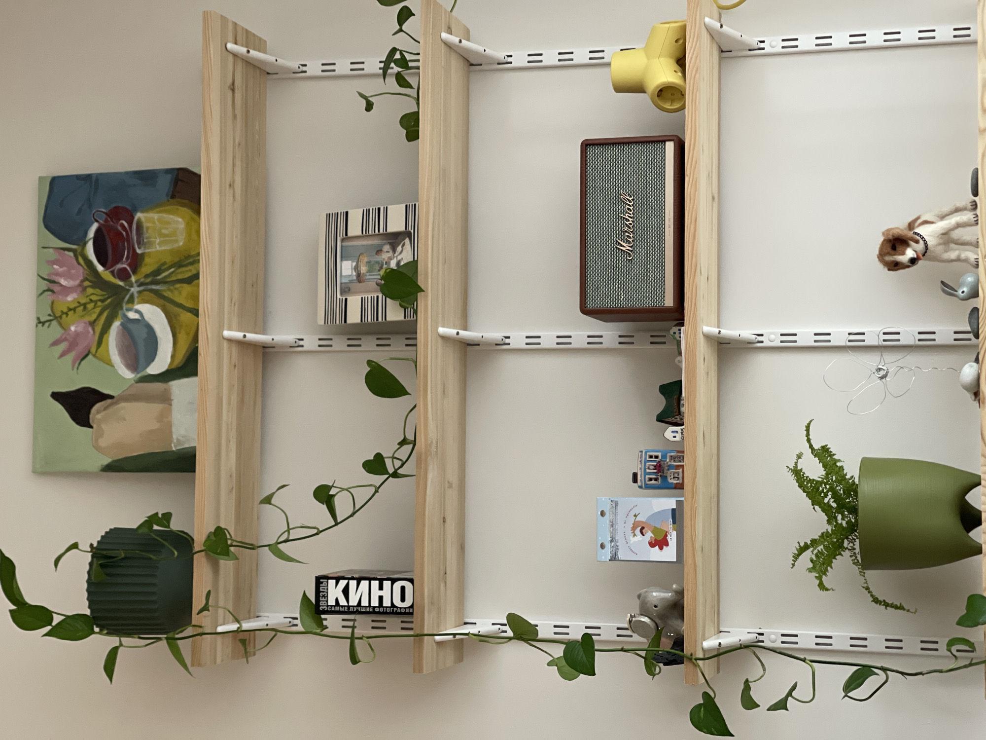 The image shows a wall-mounted shelving unit with three vertical wooden supports and horizontal metal tracks. The shelves hold a variety of objects, including a Marshall speaker, a yellow toy, a picture frame, a "KINO" book, a green textured pot, a green plant in a green pot, a toy elephant, a toy house, and a toy dog. A painting is hanging on the wall to the left of the shelves. A vine is draped around the shelves. "KINO" is printed in Cyrillic with accompanying Russian text "ЗВЕЗДЫ САМЫЕ ЛУЧШИЕ ФОТОГРАФИИ", which translates to "STARS THE BEST PHOTOS". The location, Izhevsk, Russia, is inferred from the "KINO" book.