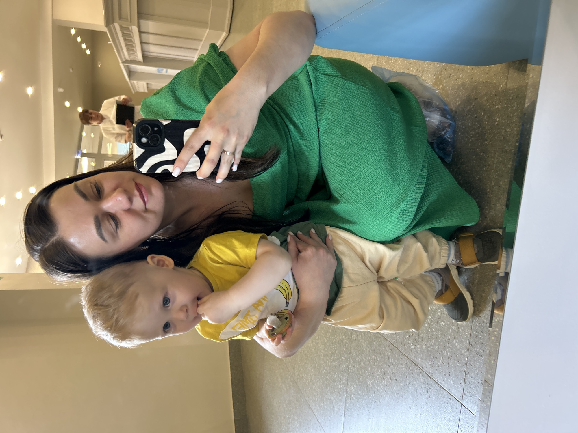 A woman in a green dress is taking a selfie with a baby in a yellow shirt in what appears to be a mirror. The baby is holding a pacifier. In the reflection, a person holding a black tablet can be seen. The scene is set in a building, with lights and interior design visible in the background. Based on the available information, determining the specific location in Izhevsk, Russia is impossible.